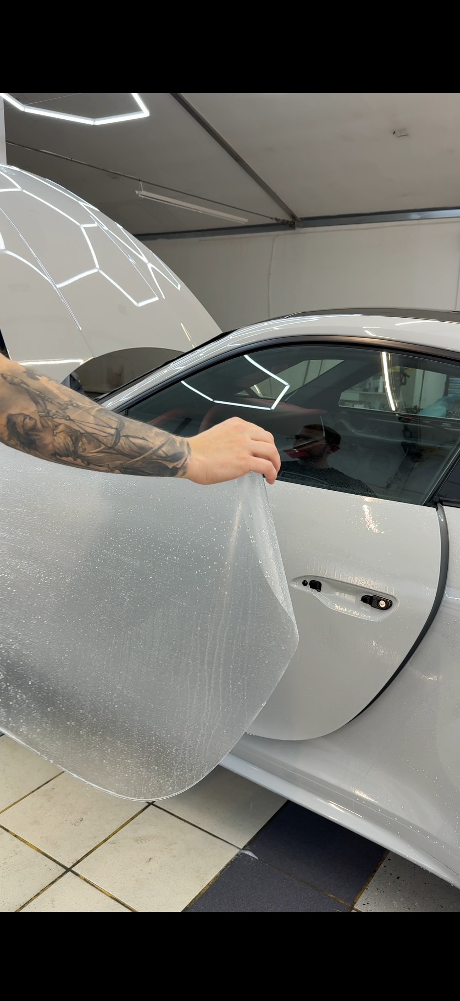 Nakładanie folii ochronnej na białe auto sportowe w dobrze oświetlonym warsztacie. Widoczna wytatuowana ręka aplikująca folię na drzwi pojazdu.