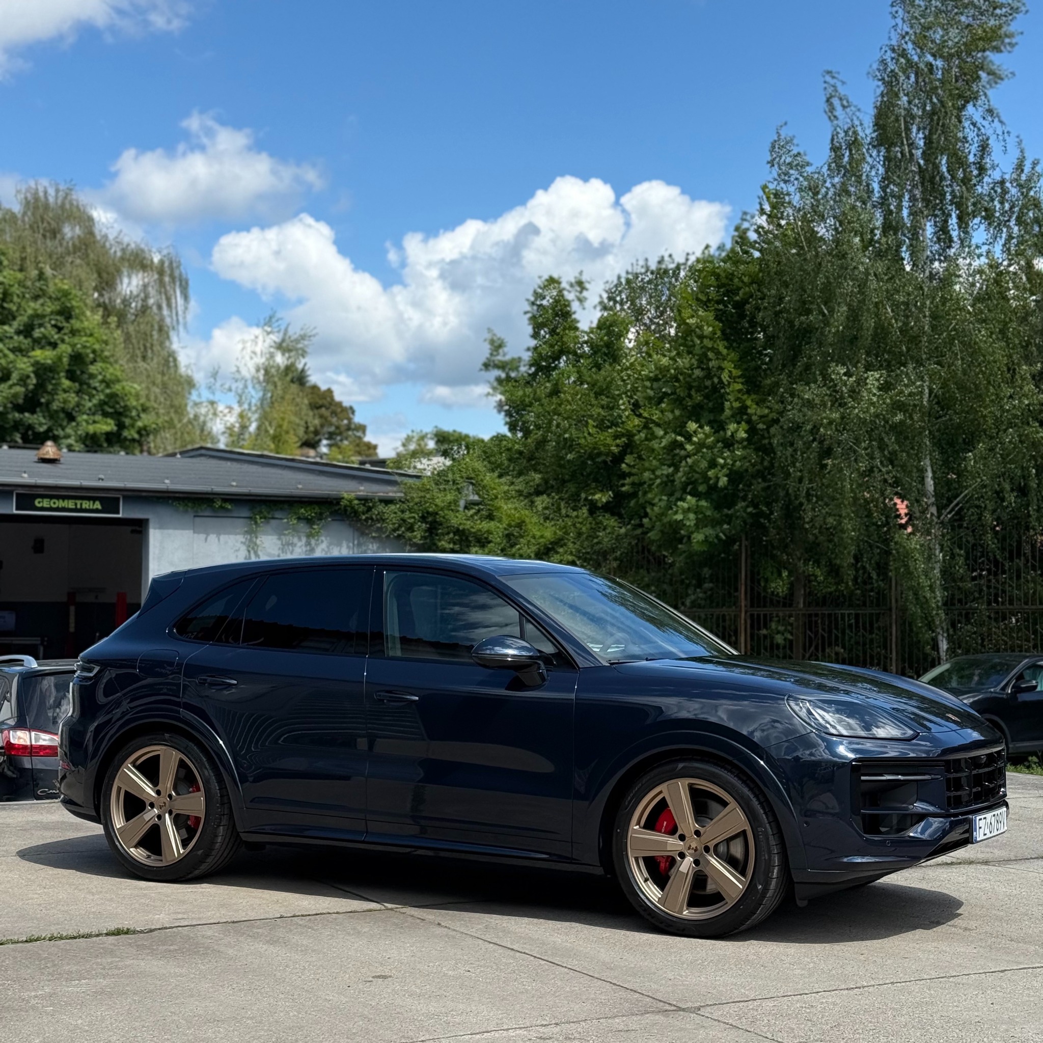 Ciemny SUV Porsche Cayenne z złotymi felgami na tle budynku z napisem 'Geometria' i zieleni. Tuning wizualny, felgi aluminiowe, opony letnie.