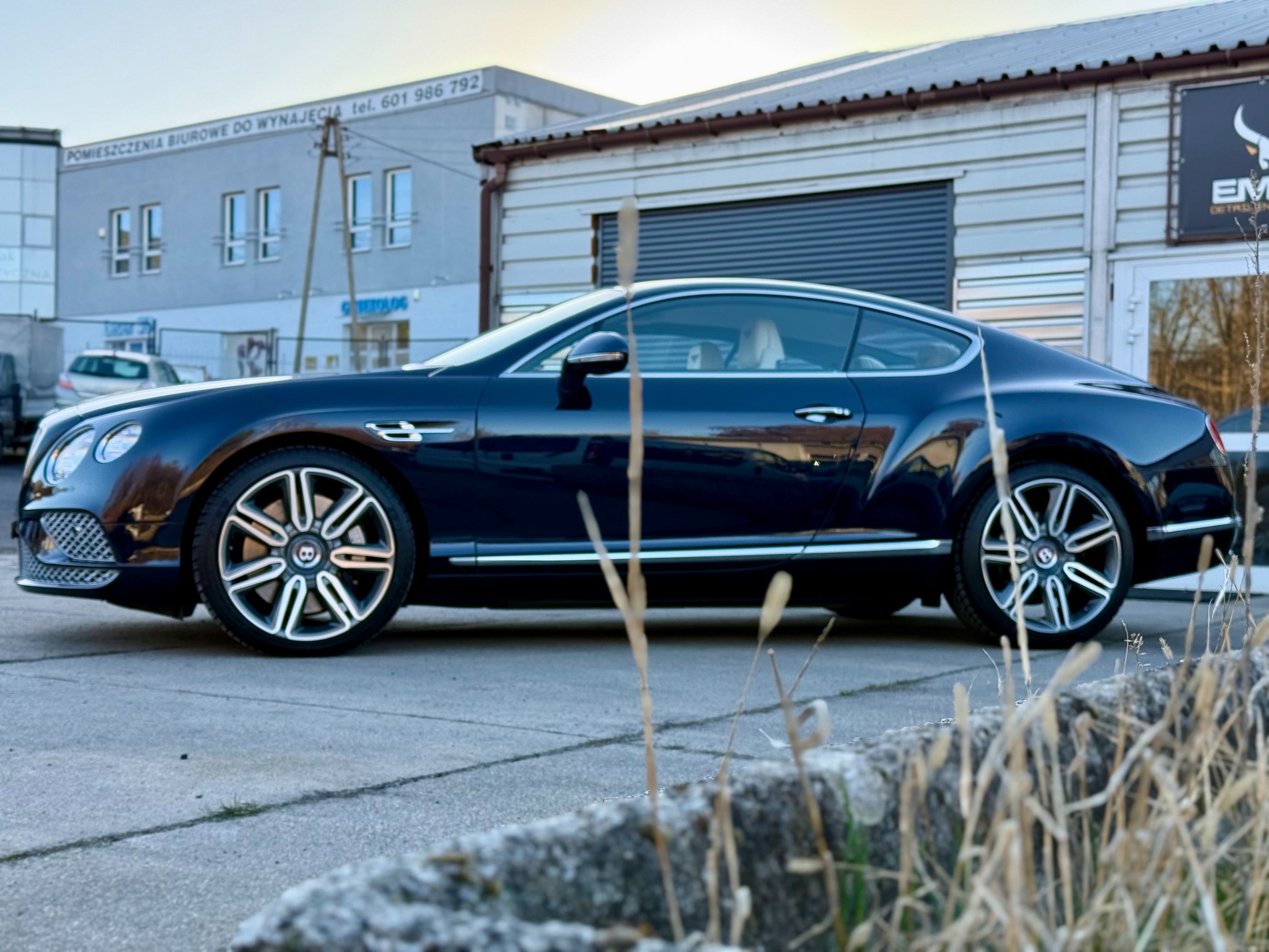 Granatowy Bentley Continental GT po detailingu, zaparkowany przed budynkiem z szarą elewacją. Widoczne logo firmy detailingowej na drzwiach. Ujęcie z niskiej perspektywy.