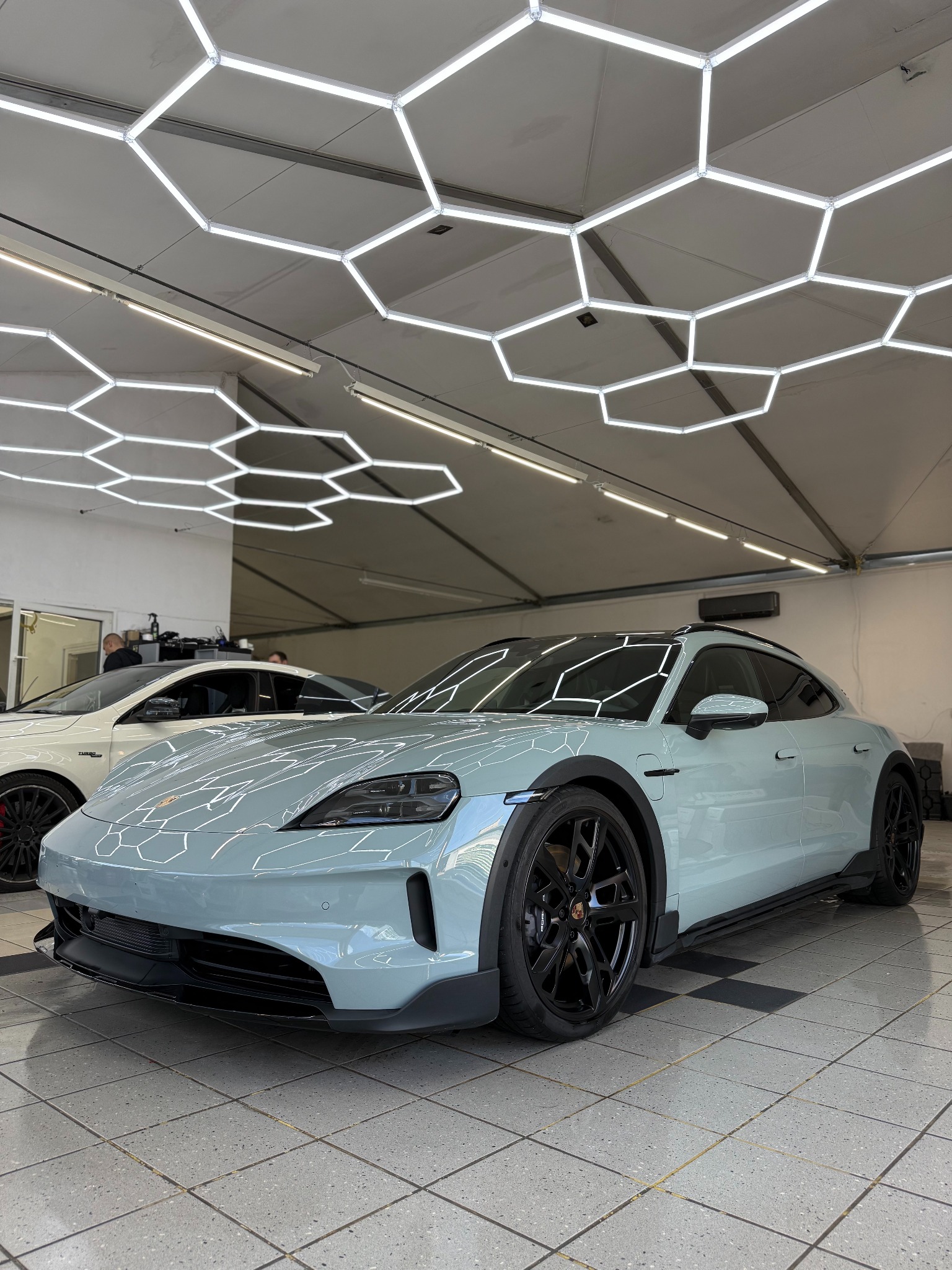 Błękitny Porsche Taycan z czarnymi felgami w jasnym pomieszczeniu z heksagonalnym oświetleniem sufitowym. Widoczny fragment innego auta w tle.