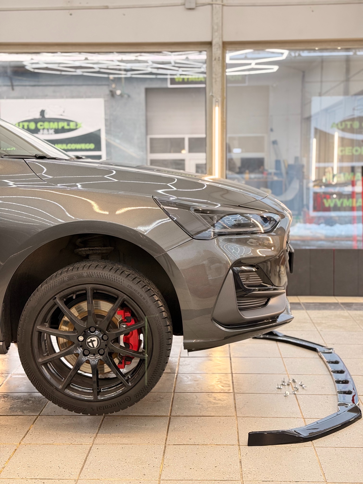 Szary samochód z czarnymi felgami i czerwonymi zaciskami hamulcowymi. Obok leży czarna dokładka zderzaka z zestawem śrub. Widok na przód auta w jasnym pomieszczeniu.