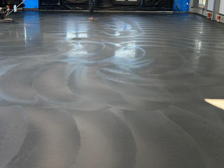 Błyszcząca, szara posadzka w pomieszczeniu po polerowaniu, z widocznymi śladami maszyny polerskiej układającymi się w łagodne fale, odbijająca światło okien.