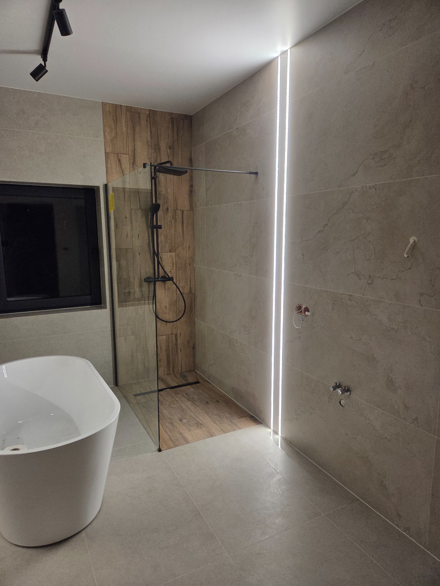 Nowoczesna łazienka z kabiną prysznicową typu walk-in, drewnopodobnymi płytkami w strefie prysznica i podświetleniem LED wzdłuż ściany. Minimalistyczna wanna wolnostojąca.