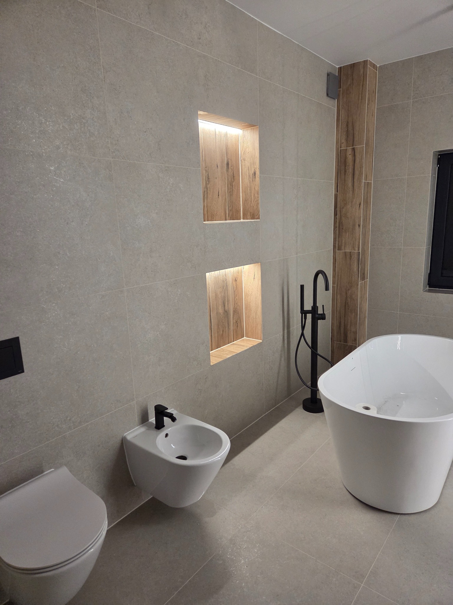 Wykończona łazienka w Starym Mieście z wanną wolnostojącą, bidetem i WC. Szare płytki, drewniane wnęki z oświetleniem. Nowoczesny design.