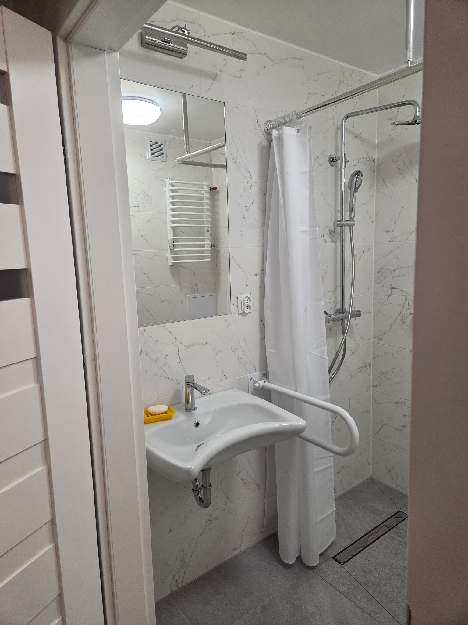 Biała łazienka z marmurowymi płytkami, umywalką z uchwytem dla niepełnosprawnych i kabiną prysznicową z zasłoną. Widoczny odpływ liniowy i grzejnik drabinkowy.