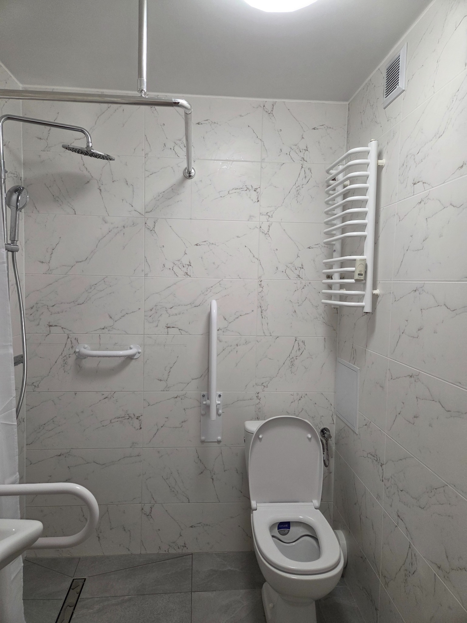 Jasna łazienka z marmurowymi płytkami, wyposażona w uchwyty dla niepełnosprawnych, grzejnik drabinkowy i kabinę prysznicową z deszczownicą. Uwagę zwraca toaleta z deską sedesową.