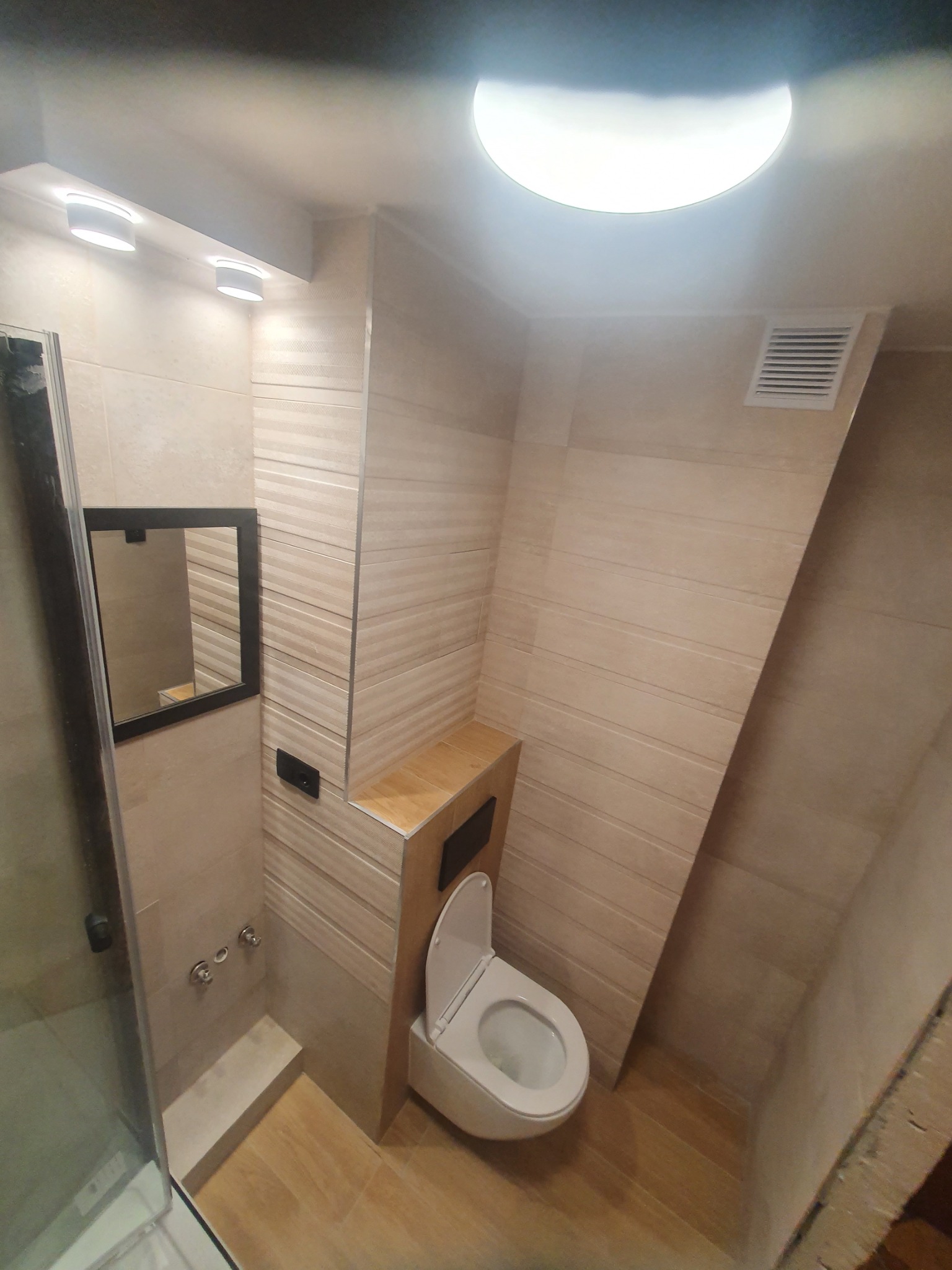 Wykończona łazienka z beżowymi płytkami imitującymi drewno, podwieszana toaleta z drewnianą półką, czarne lustro w ramie i przeszklony prysznic.