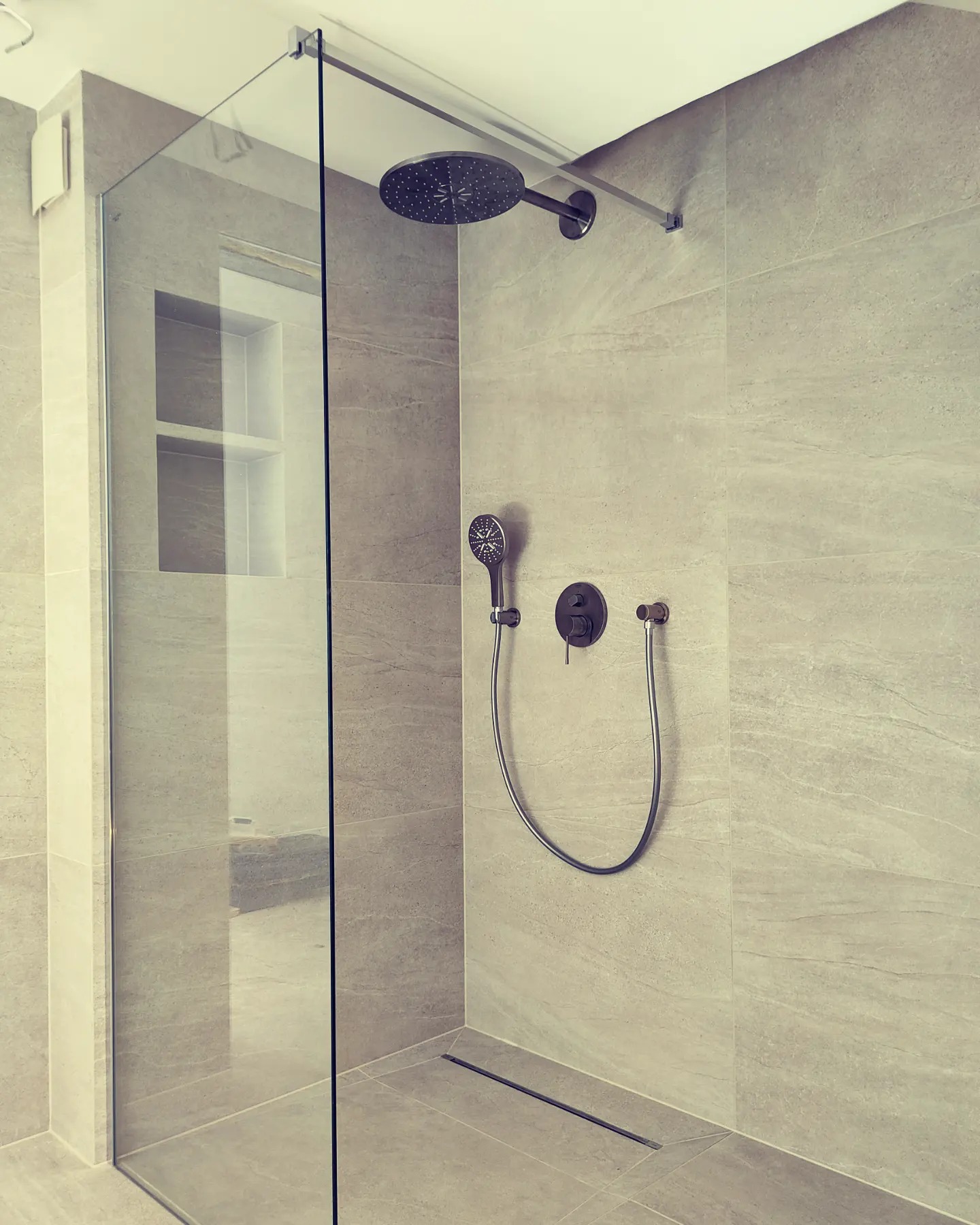 Nowoczesna kabina prysznicowa typu walk-in z deszczownicą i baterią podtynkową w łazience wykończonej płytkami imitującymi kamień. Widoczny odpływ liniowy w podłodze.