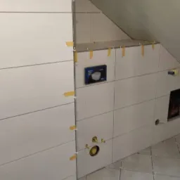 Białe płytki przyklejane do ściany w łazience na poddaszu, przytrzymywane żółtą taśmą, widoczna instalacja wodna i pojemnik z narzędziami i odpadami.