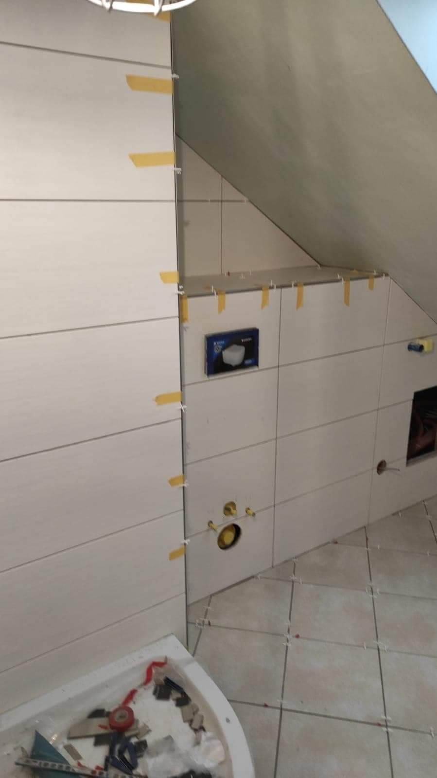 Białe płytki przyklejane do ściany w łazience na poddaszu, przytrzymywane żółtą taśmą, widoczna instalacja wodna i pojemnik z narzędziami i odpadami.