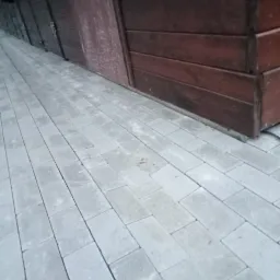 Ułożona szara kostka brukowa przy drewnianej ścianie budynku z widocznymi deskami, ułożona pod kątem, widok z niskiej perspektywy.