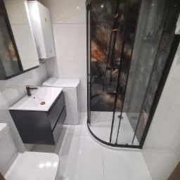 Nowoczesna łazienka z kabiną prysznicową z czarną ramą, umywalką z szafką w kolorze antracytowym, pralką i białymi płytkami na ścianach i podłodze.