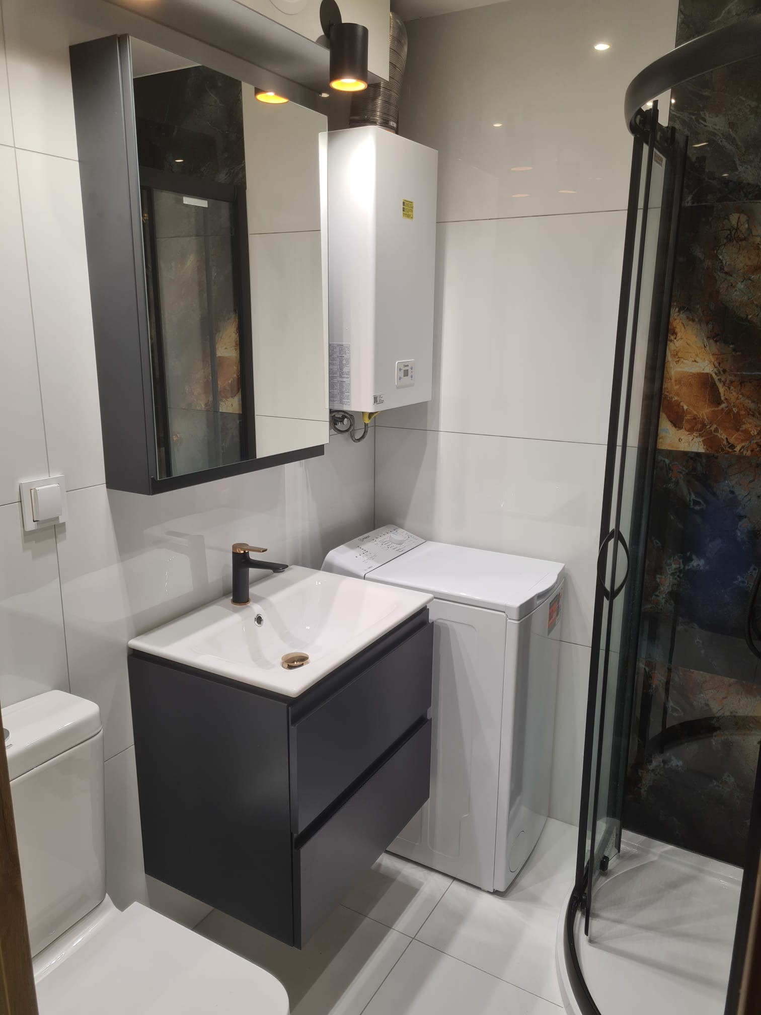 Nowoczesna łazienka po remoncie: umywalka z szafką w kolorze grafitowym, bateria w kolorze szczotkowanego złota, pralka i kabina prysznicowa z ciemnymi profilami na tle białych płytek.