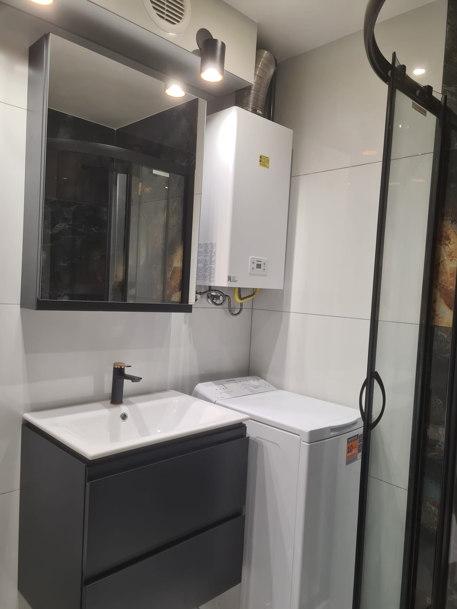 Nowoczesna łazienka z umywalką nablatową na szafce w kolorze antracytu, pralką i piecem gazowym na tle białych płytek.