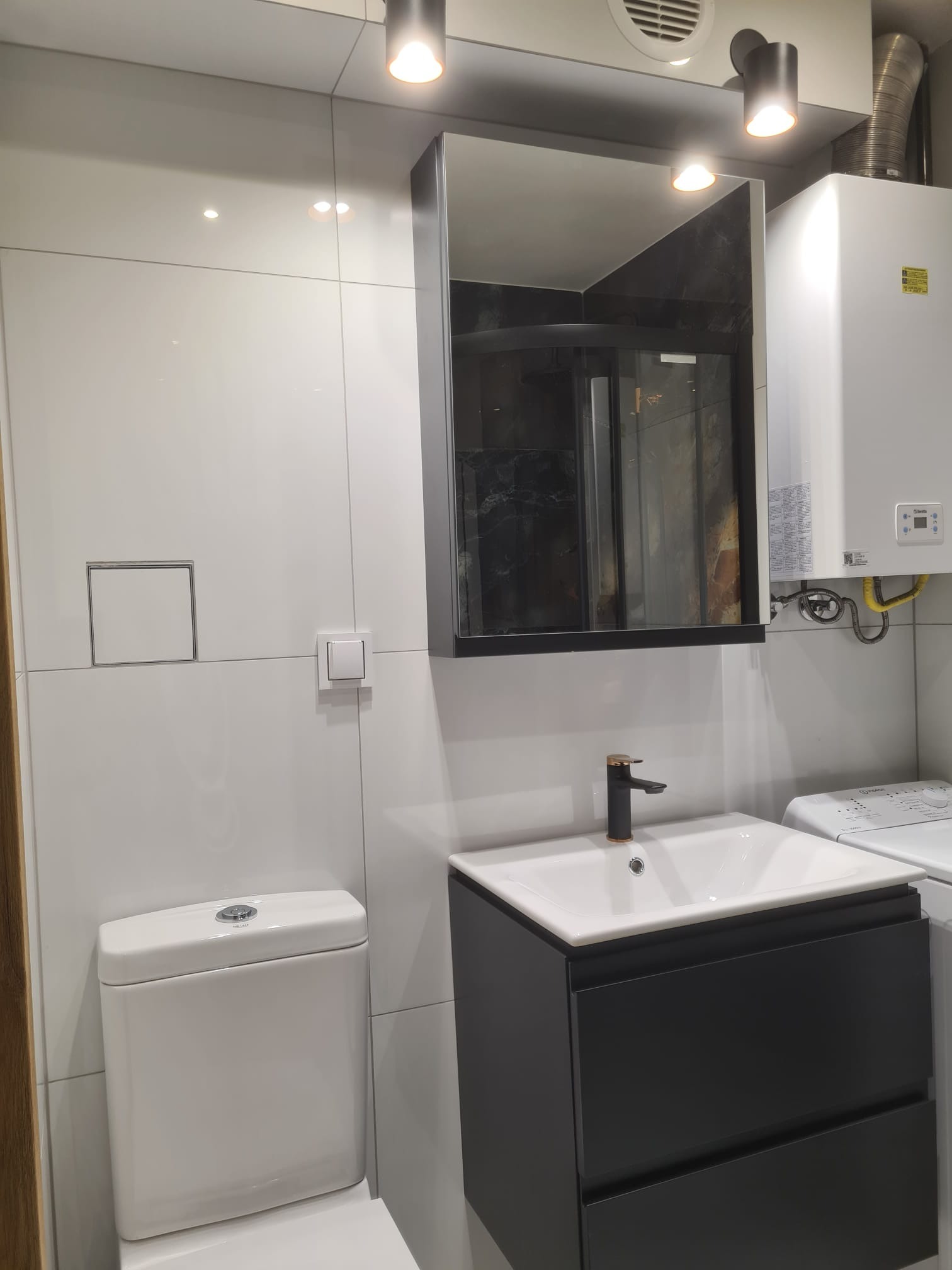 Nowoczesna łazienka z białymi płytkami, wiszącą toaletą, umywalką z czarną szafką i złotą baterią oraz lustrem w ciemnej ramie, oświetlona punktowymi lampami.