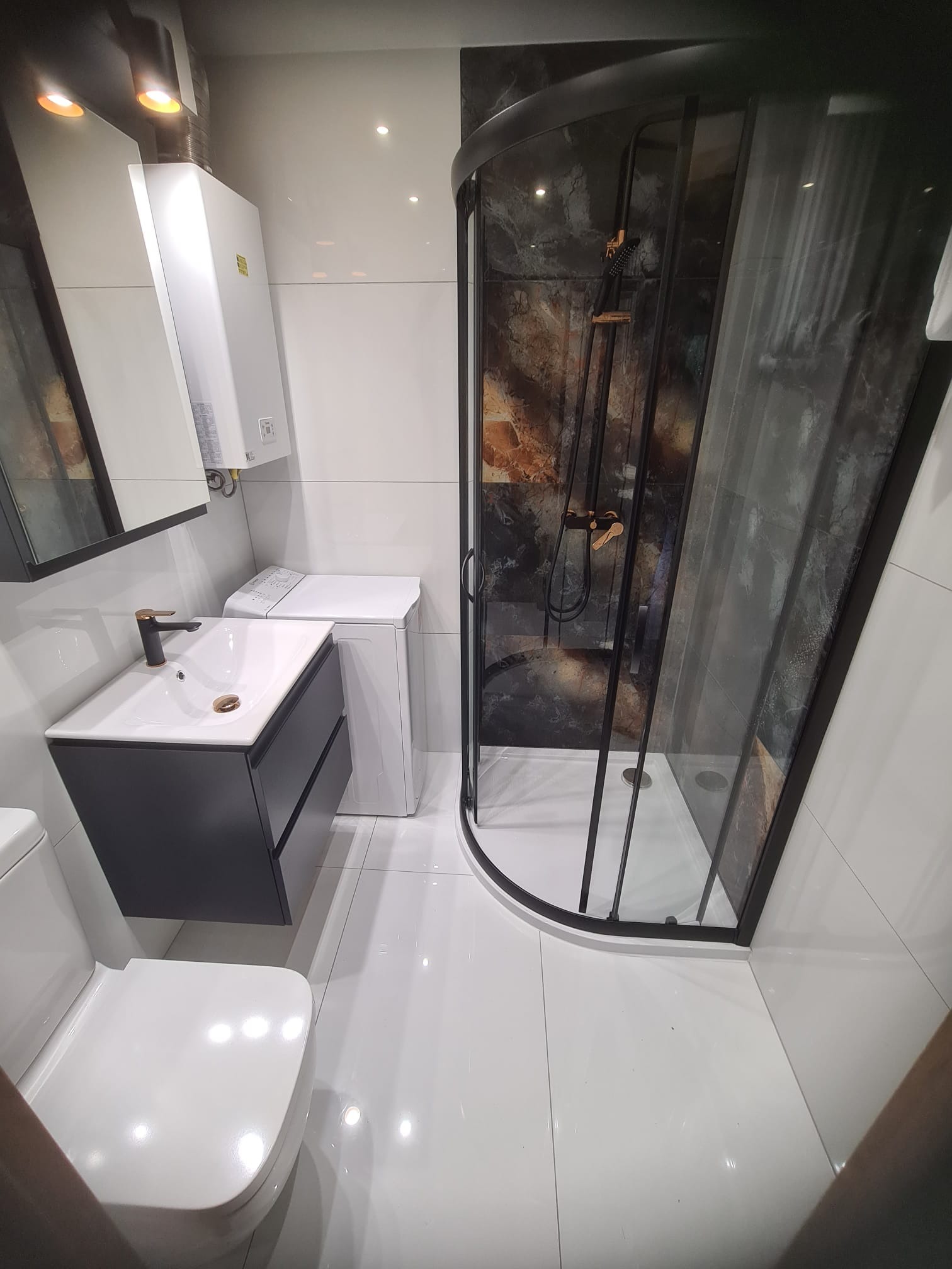 Nowoczesna łazienka z kabiną prysznicową z czarną ramą, umywalką z szafką w kolorze antracytowym, pralką i białymi płytkami na ścianach i podłodze.