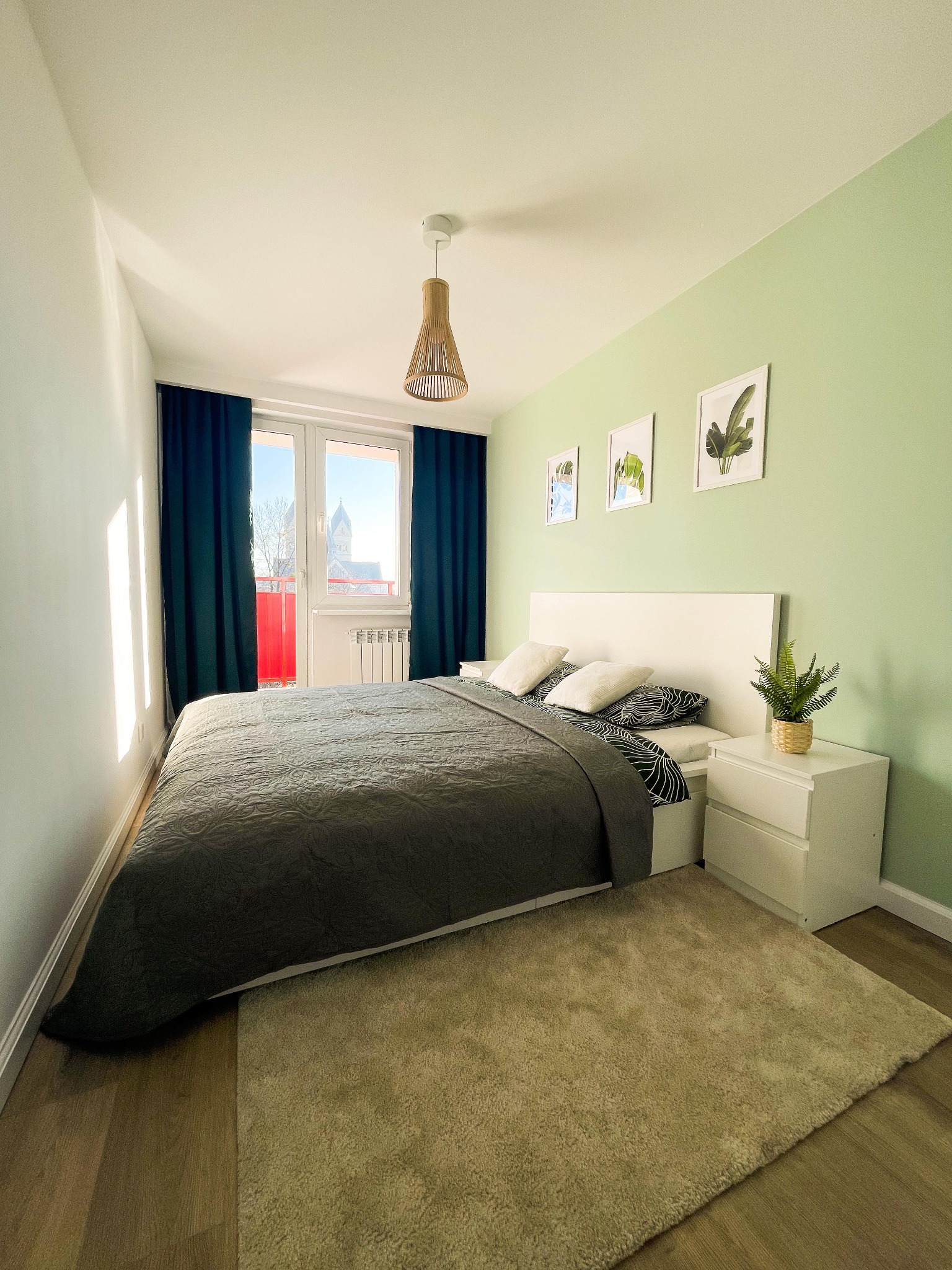 Jasna sypialnia z łóżkiem, drewnianą podłogą, zieloną ścianą i widokiem na kościół za oknem.