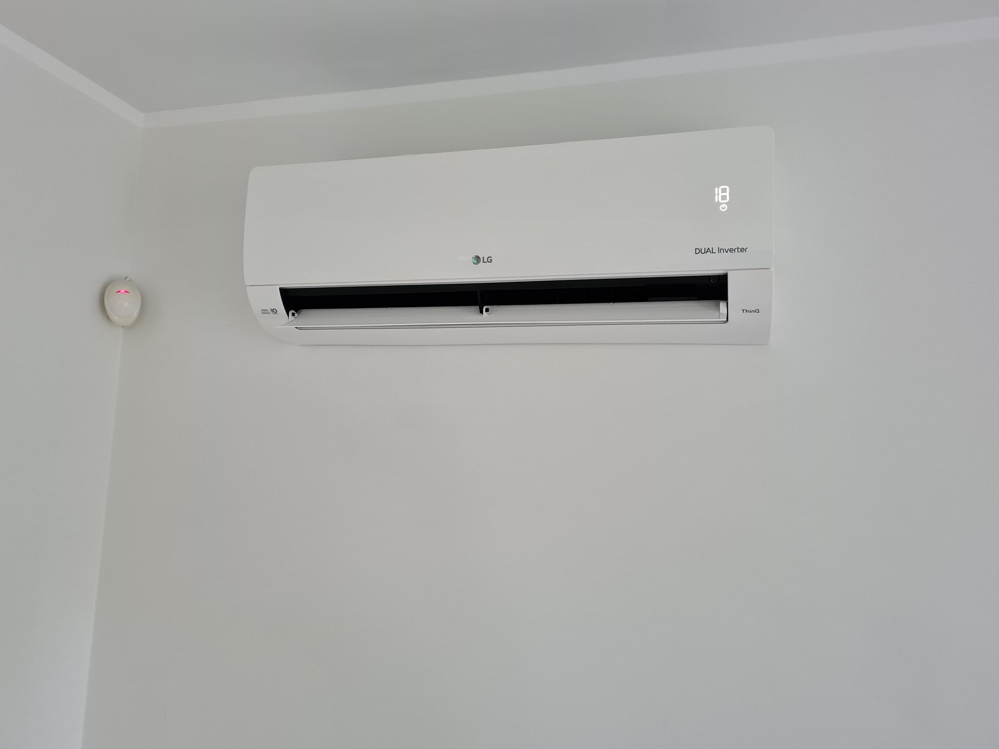 Biały klimatyzator LG Dual Inverter zamontowany na jasnej ścianie, widoczny czujnik dymu w rogu pomieszczenia.
