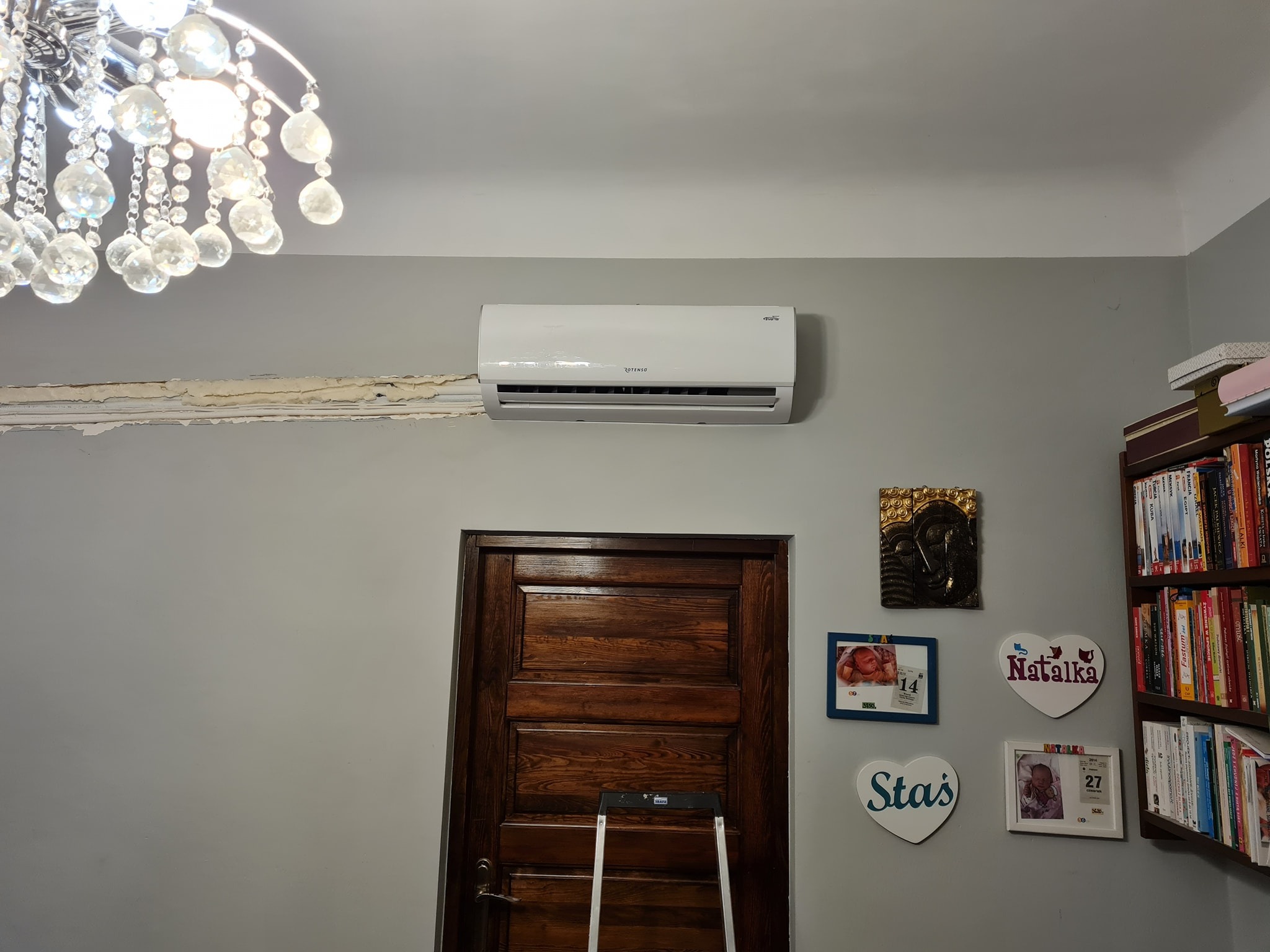 Biały klimatyzator Rotenso zamontowany na szarej ścianie nad drewnianymi drzwiami, widoczne ślady po wcześniejszym montażu, kryształowy żyrandol u góry po lewej, książki na półce po prawej...