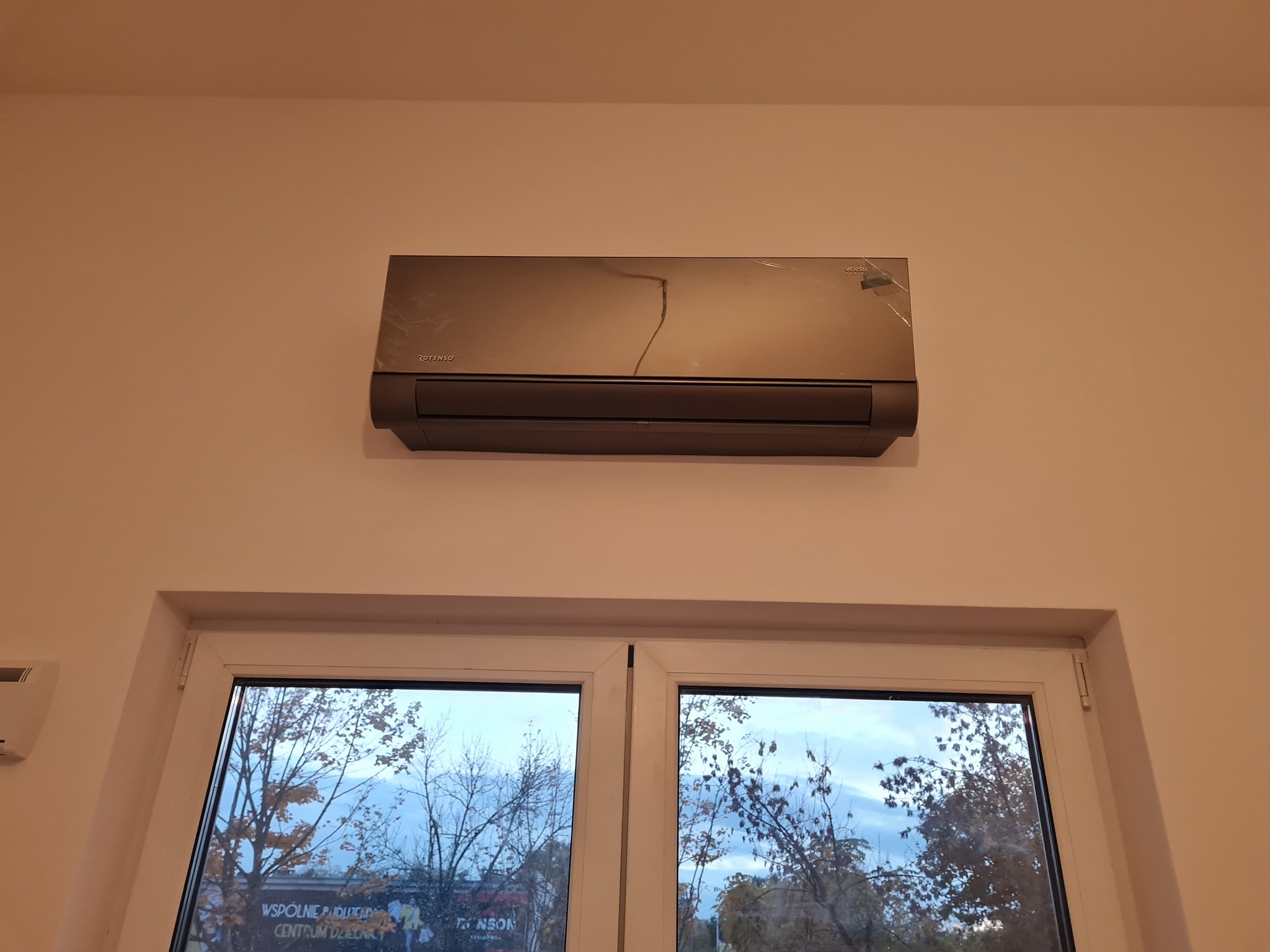 Szary klimatyzator Rotenso Versu zamontowany na ścianie nad białym oknem, widoczne drzewa za oknem