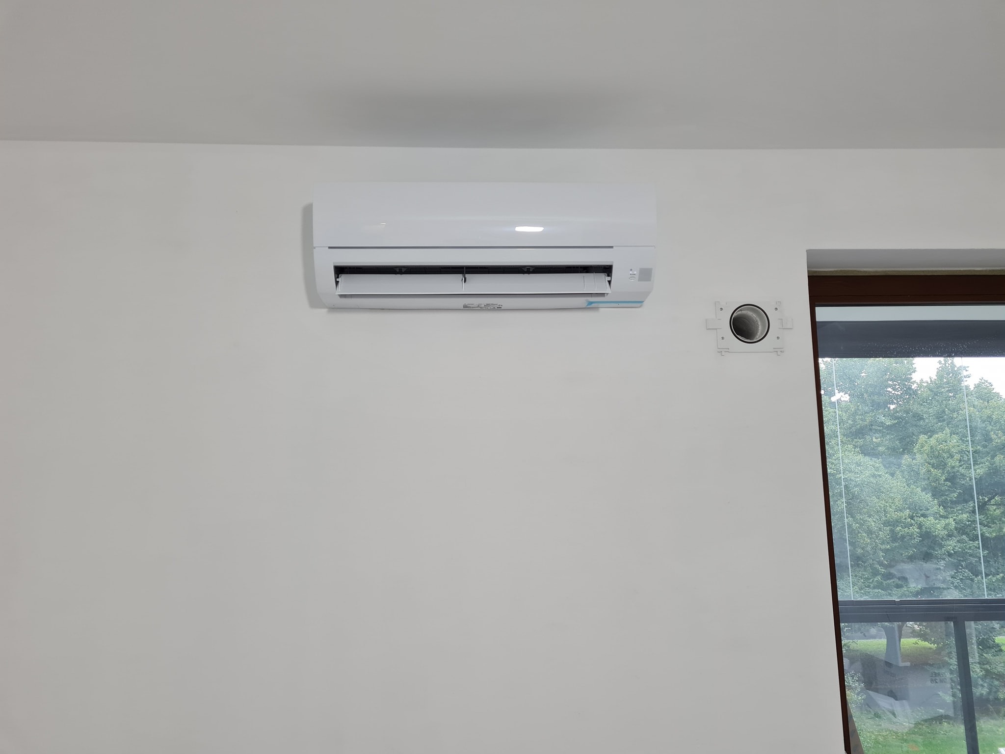 Biały klimatyzator ścienny zamontowany w jasnym pomieszczeniu z widokiem na zewnątrz przez okno, widoczny otwór wentylacyjny obok klimatyzatora.