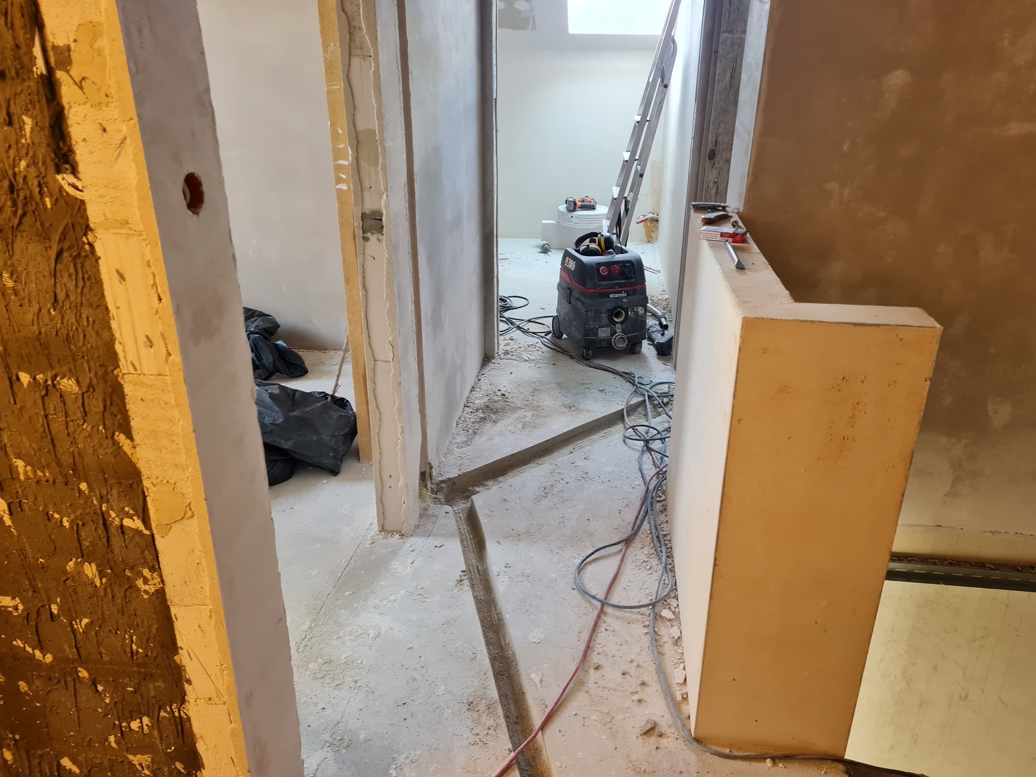 Wnętrze w trakcie remontu, widoczne kable elektryczne w bruździe w podłodze, odkurzacz przemysłowy Starmix, drabina oparta o ścianę, ściany w stanie surowym, przygotowane do wykończenia.