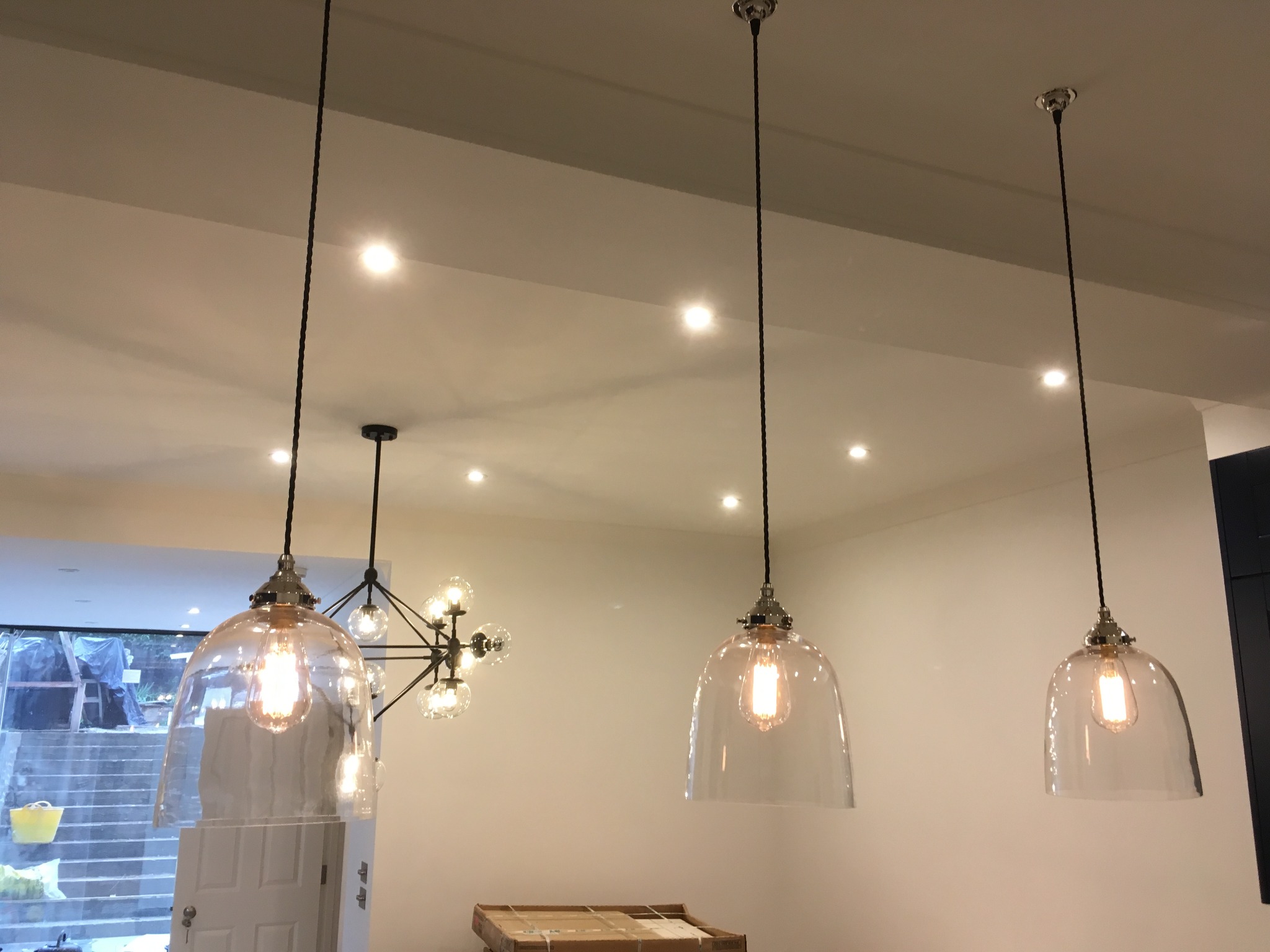 Trzy szklane lampy wiszące z żarówkami Edisona, oświetlające wnętrze z widocznym oknem i schodami w tle, oraz nowoczesny żyrandol.