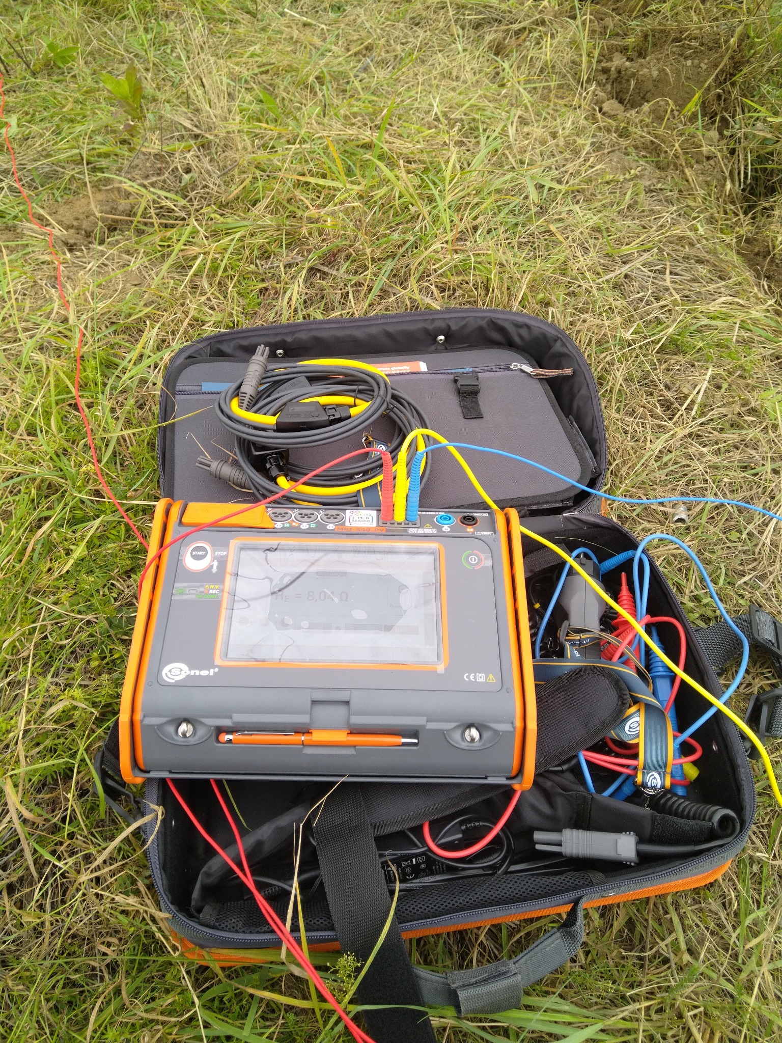 Miernik rezystancji uziomu Sonel, z widocznymi przewodami pomiarowymi rozłożonymi na trawie, w otwartej torbie transportowej.