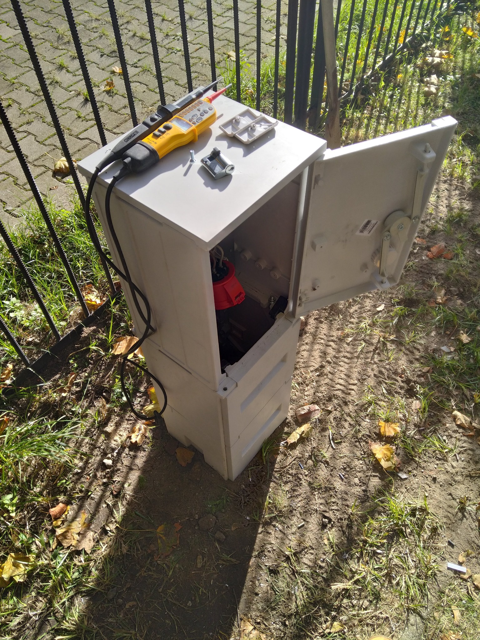 Otwarta skrzynka elektryczna na zewnątrz, z widocznym gniazdem przemysłowym w środku, obok leżą tester napięcia Fluke i elementy montażowe.