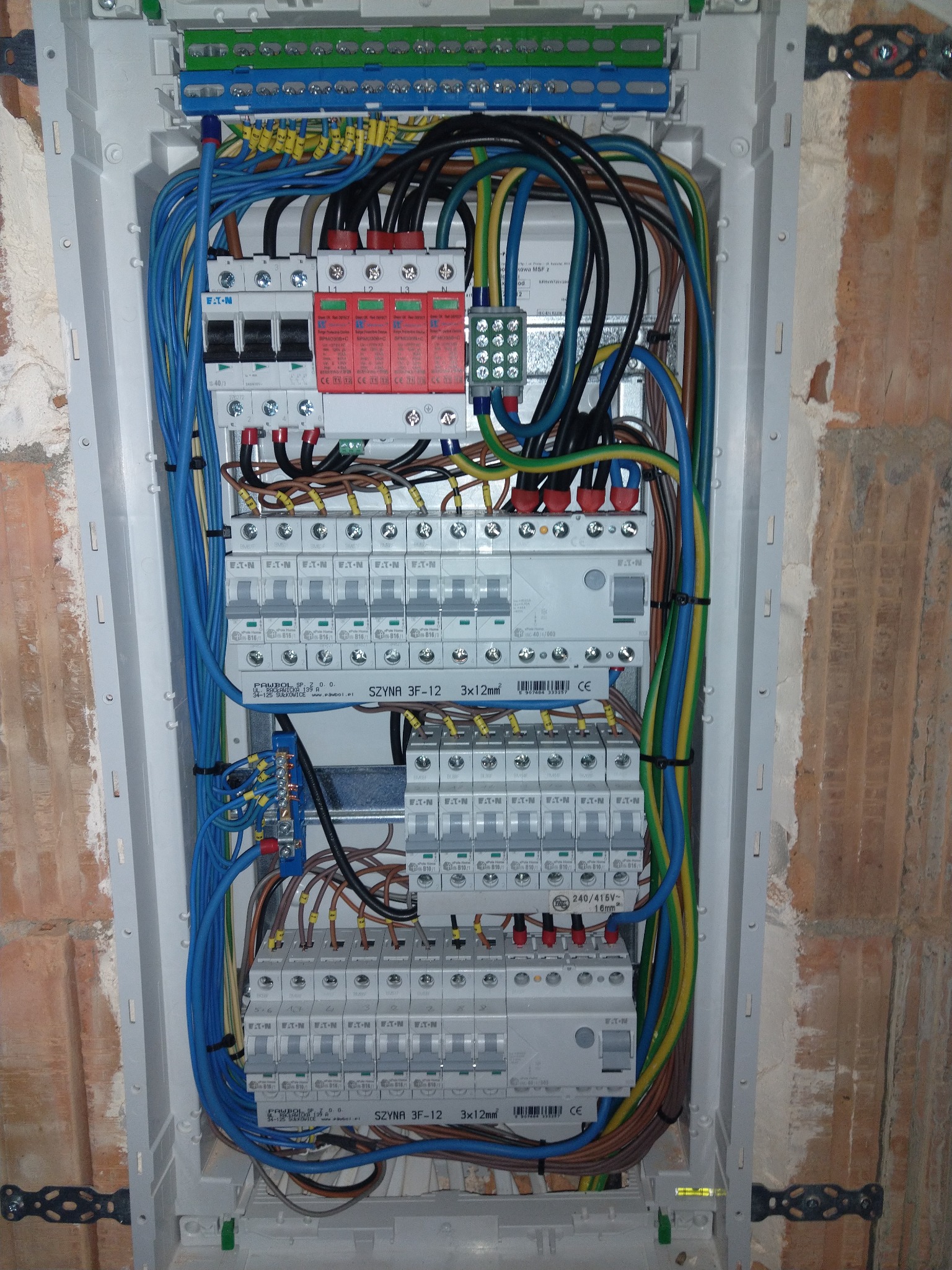 Wewnętrzny widok rozdzielnicy elektrycznej w trakcie montażu, z widocznymi przewodami w różnych kolorach, zabezpieczeniami Eaton i listwami zaciskowymi Pawbol, zamontowanej w surowej ścianie z cegły.