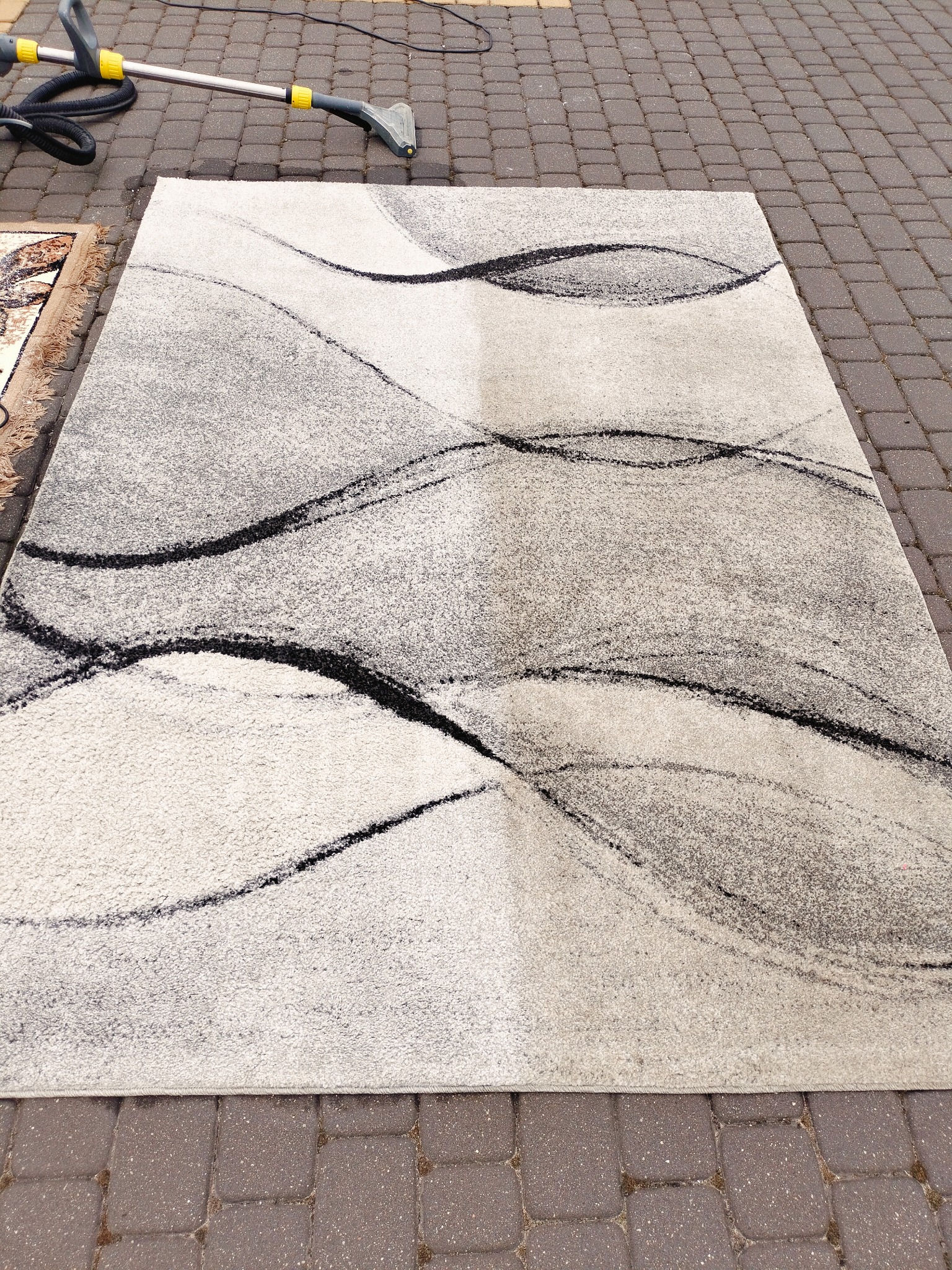 Szary dywan z abstrakcyjnym wzorem, częściowo wyczyszczony, leżący na kostce brukowej, obok widoczny sprzęt do czyszczenia.