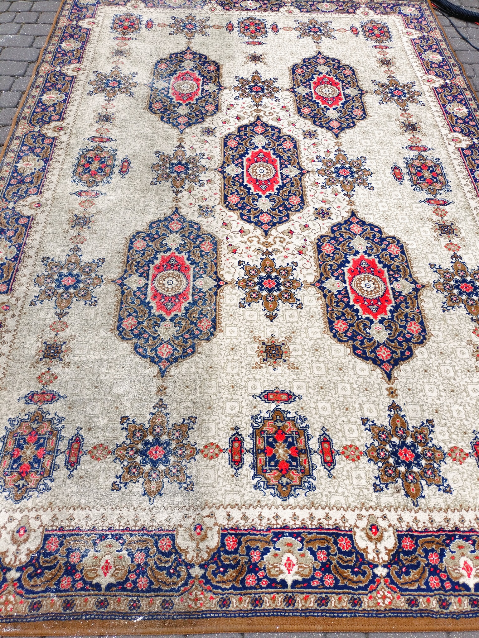 Wielkoformatowy dywan z orientalnym wzorem w kolorach czerwonym, niebieskim i brązowym, leżący na szarej kostce brukowej. Widoczne ślady po czyszczeniu parowym.