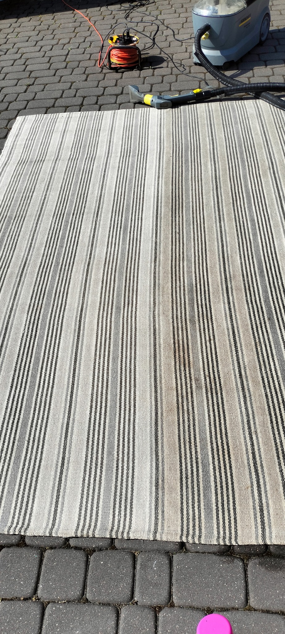 Pasiasty dywan z widocznymi plamami, leżący na kostce brukowej, obok sprzętu do czyszczenia z odciągniętym kablem.