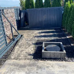 Nowoczesne ogrodzenie panelowe z gabionem wypełnionym kamieniem, brama wjazdowa i fragment podjazdu z kostki brukowej w odcieniach szarości, z elementami dekoracyjnymi z czarnego grysu.
