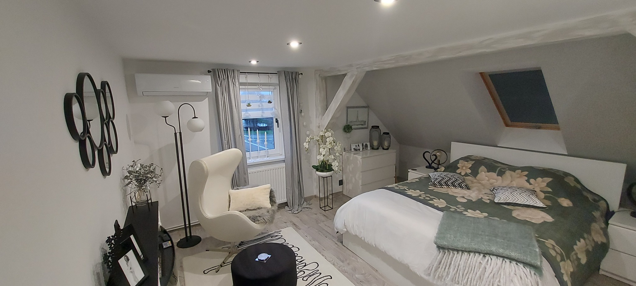 Jasne wnętrze sypialni na poddaszu z klimatyzatorem ściennym, fotelem typu Egg, białymi meblami i dekoracjami w odcieniach szarości, bieli i czerni.