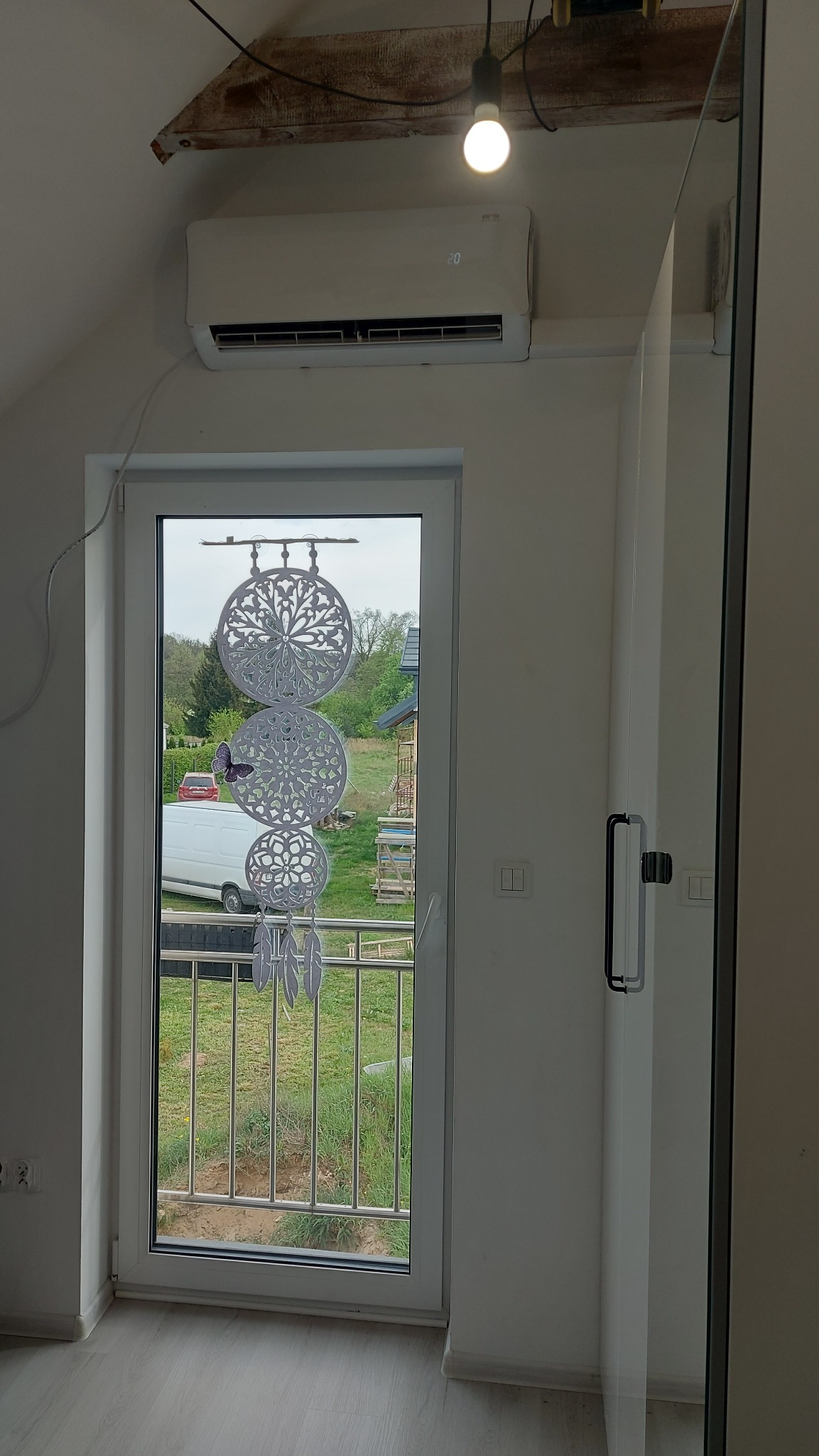 Biały klimatyzator ścienny zamontowany nad oknem z widokiem na balkon i zieleń, w pokoju z drewnianą belką stropową i wiszącą żarówką.