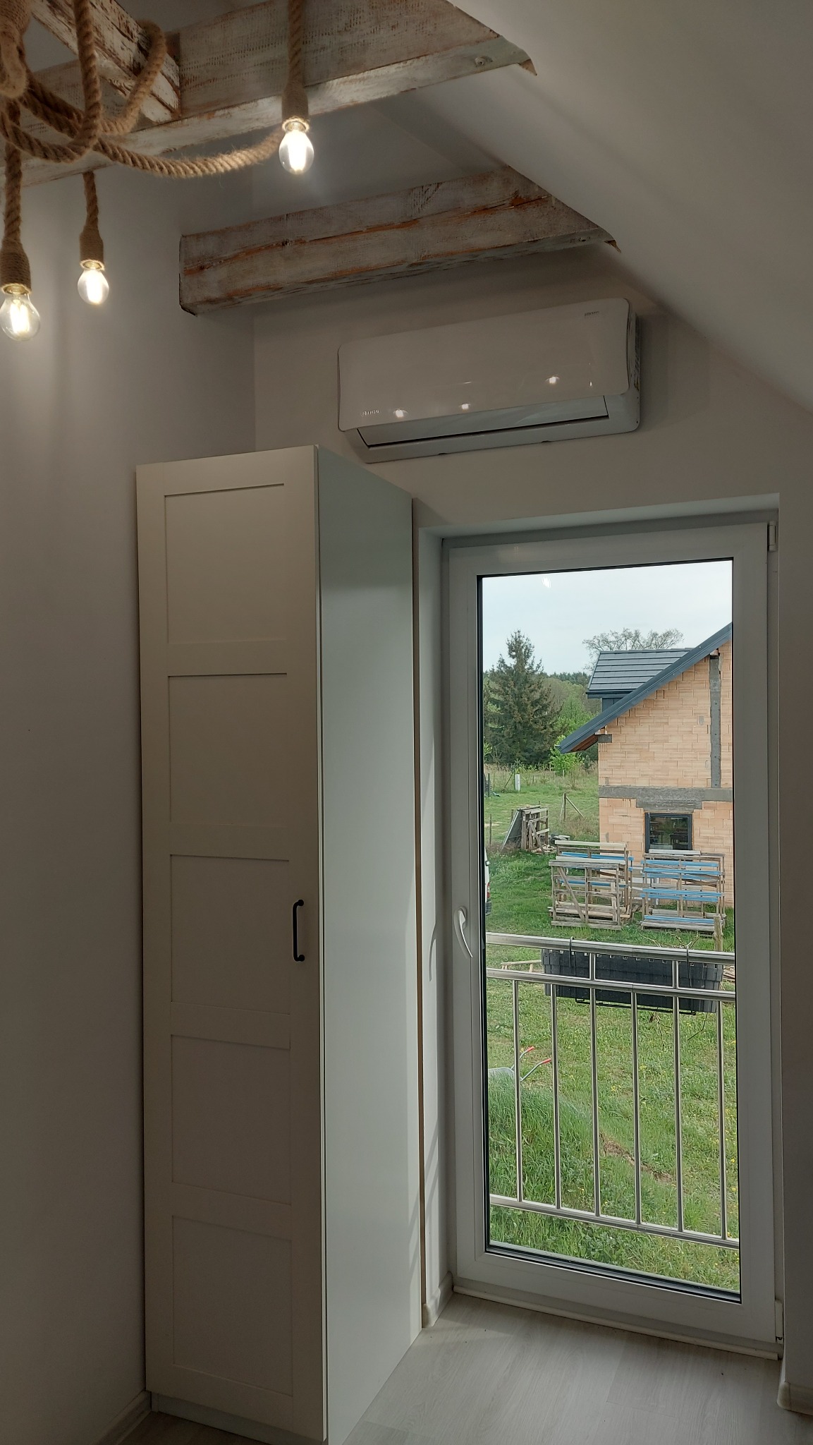 Biały klimatyzator ścienny zamontowany nad szafą w pokoju z oknem na ogród, dekoracyjne oświetlenie z linami i żarówkami.