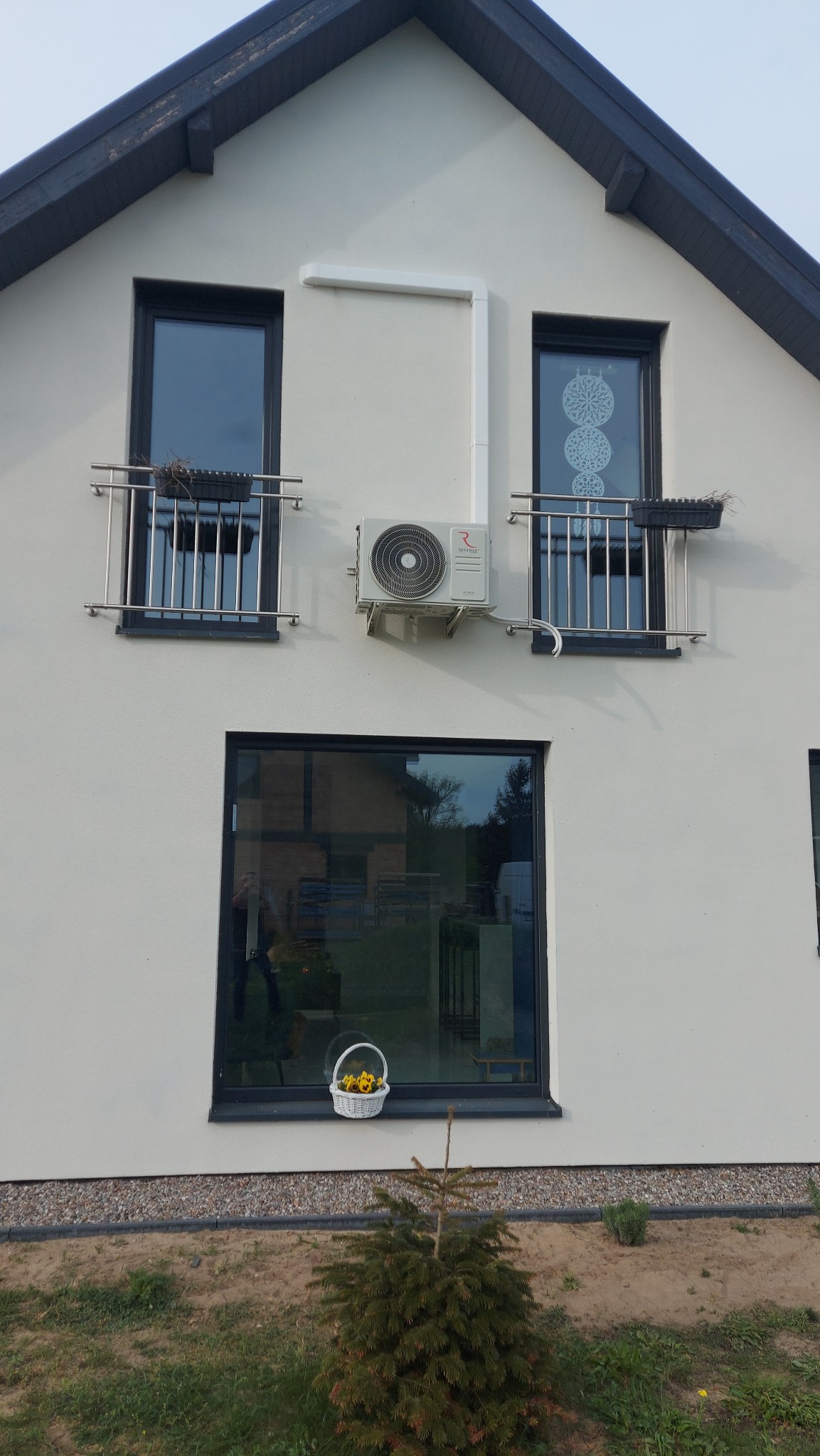 Biały dom jednorodzinny z zamontowaną klimatyzacją Rotenso, widoczne rury odprowadzające, okna z balkonami i koszykiem z kwiatami na parapecie.