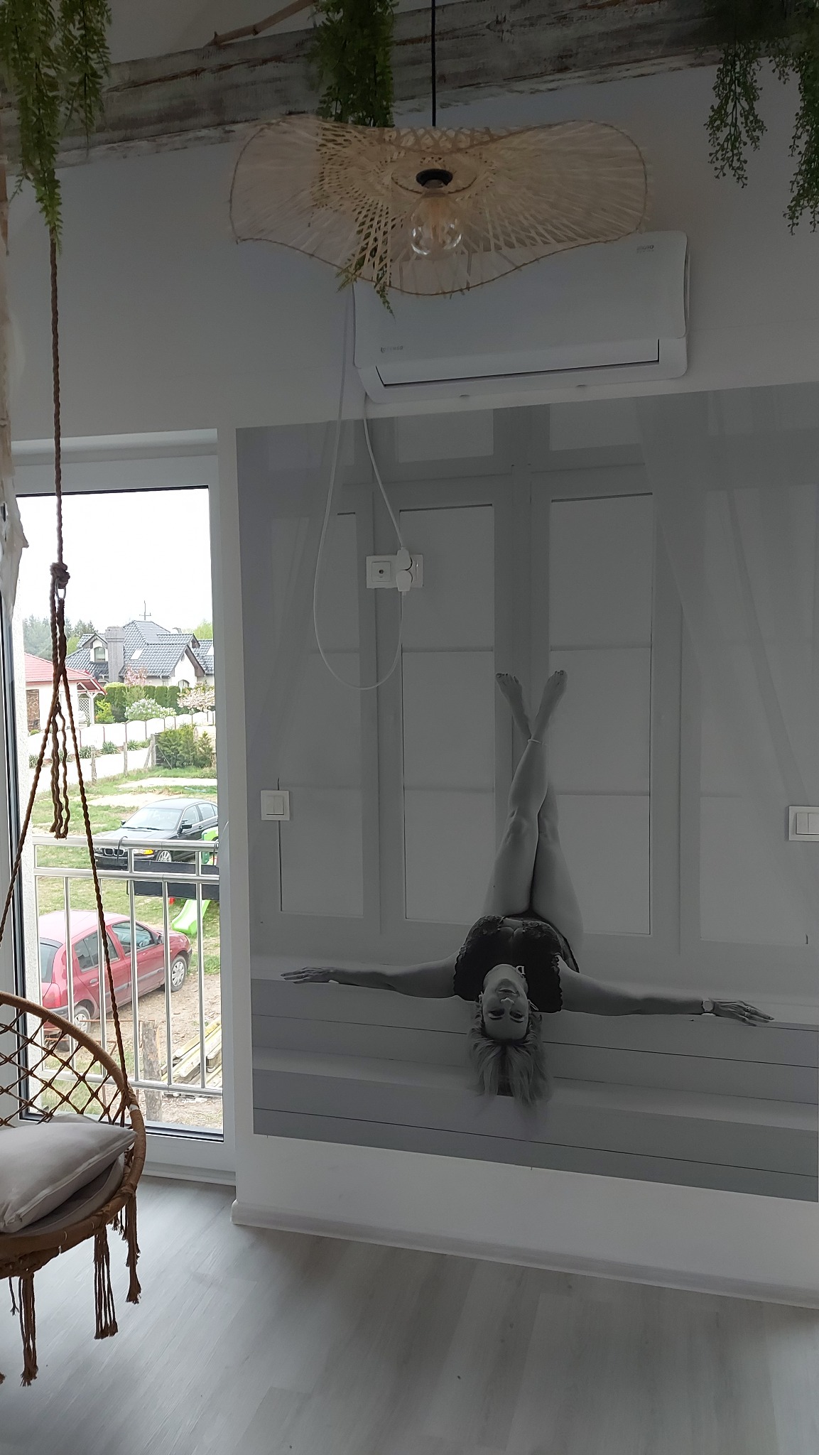 Biały klimatyzator ścienny zamontowany w pomieszczeniu z dekoracyjną lampą z naturalnego materiału i fototapetą przedstawiającą osobę w pozycji odwróconej.