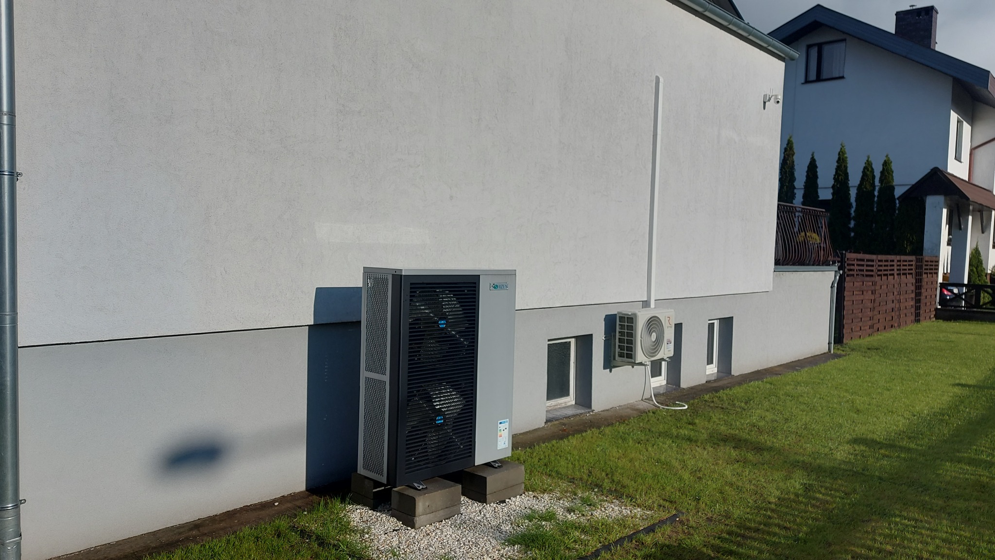Zewnętrzna jednostka klimatyzacji typu split zamontowana na betonowych bloczkach przy ścianie domu, z widoczną drugą mniejszą jednostką na ścianie i fragmentem trawnika.