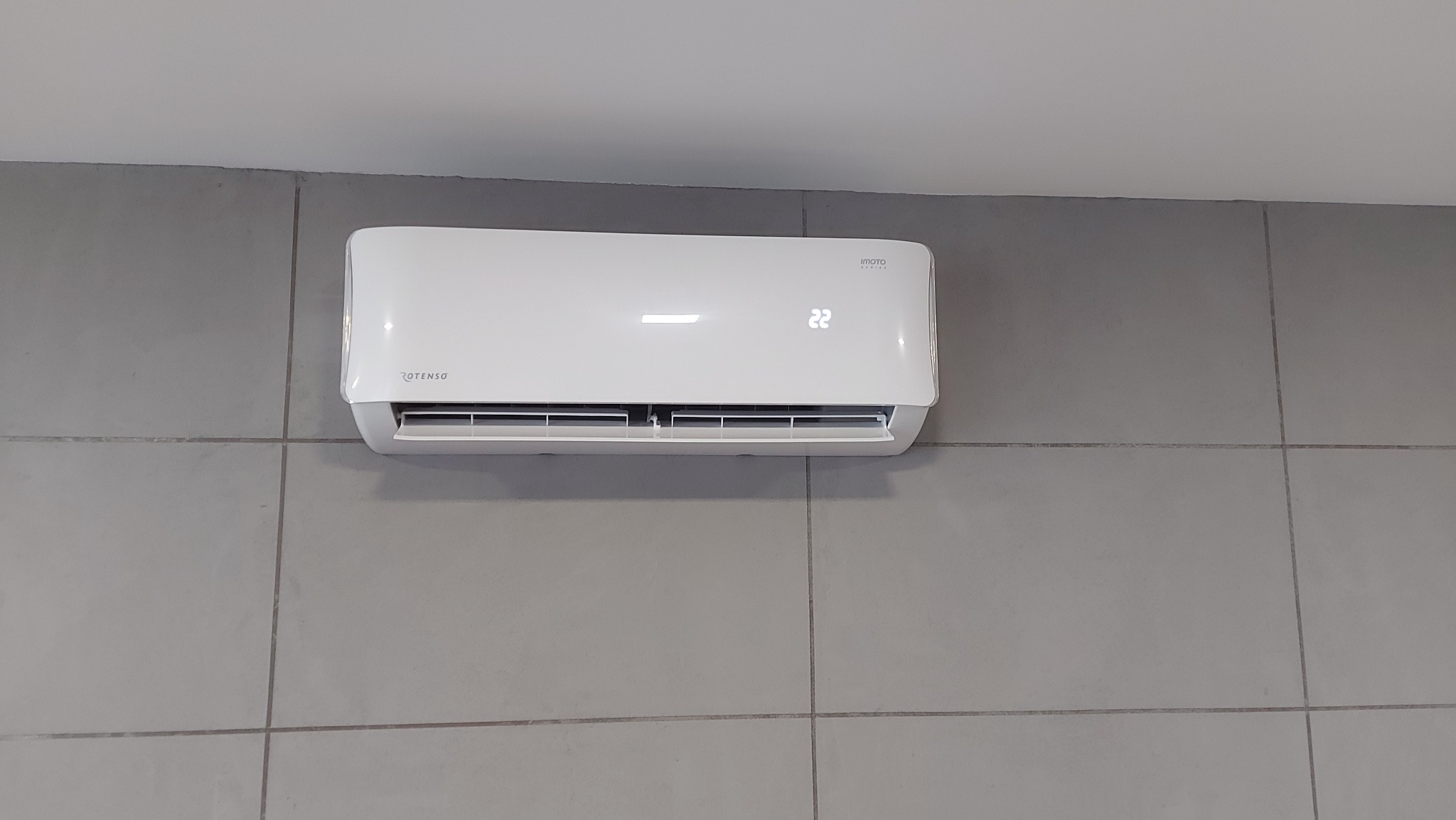 Biały klimatyzator Rotenso z wyświetlaczem temperatury 22 stopnie, zamontowany na szarej, kafelkowanej ścianie.