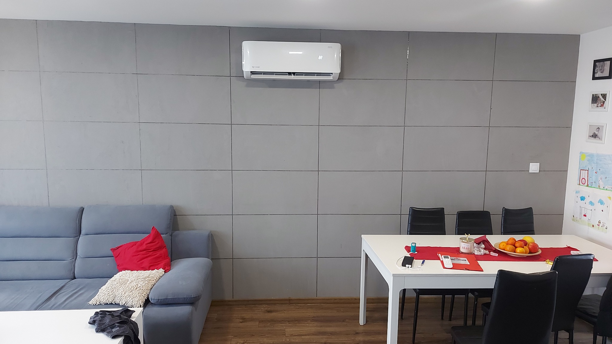 Biały klimatyzator ścienny w szarym salonie nad szarą sofą i białym stołem jadalnym.