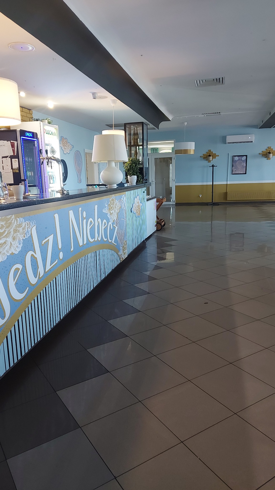 Wnętrze lokalu w Międzyrzeczu z klimatyzatorem ściennym widocznym w tle, nad barem z napisem 'Jedz! Niebecz'.
