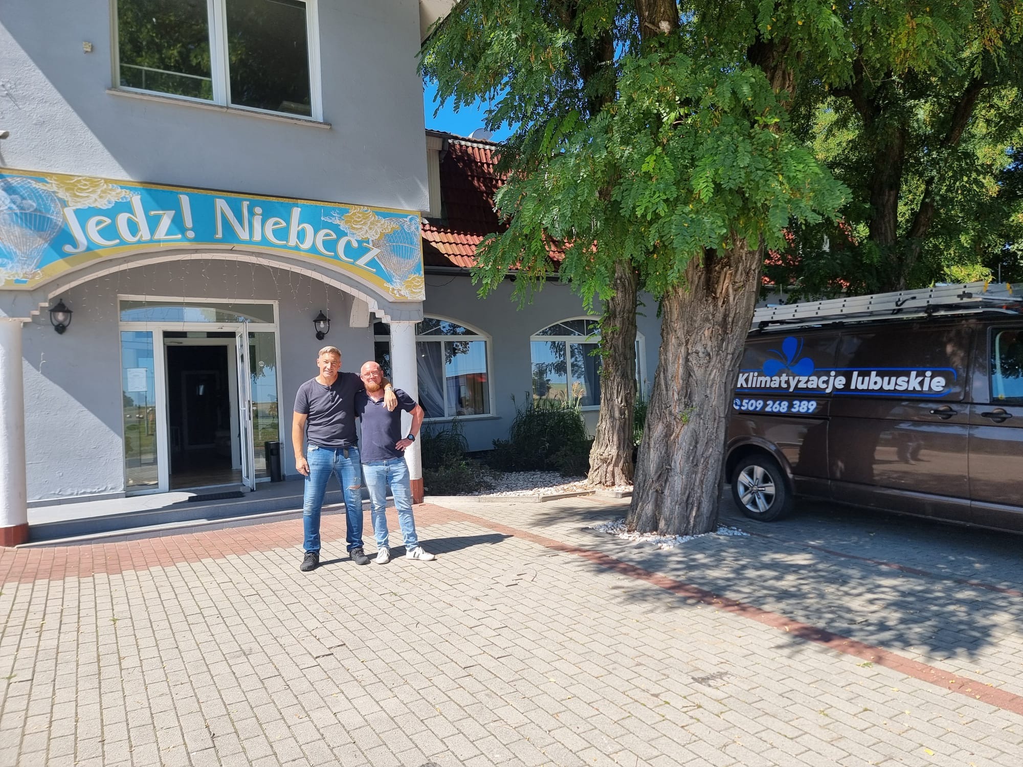Dwaj mężczyźni stojący przed budynkiem z napisem 'Jedź! Niebecz' i samochodem firmy Klimatyzacje Lubuskie z drabiną na dachu.