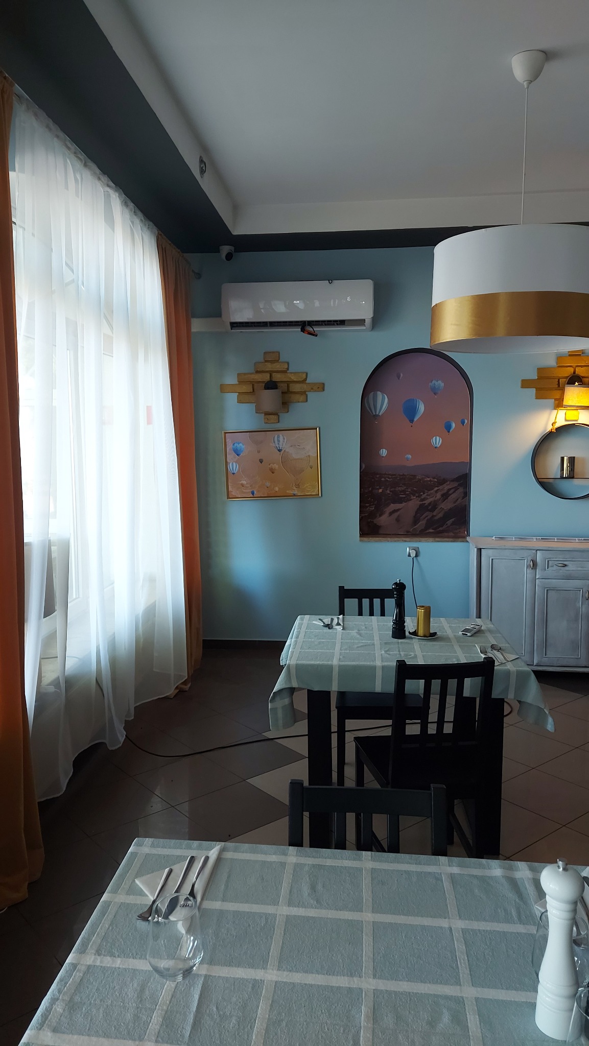 Wnętrze restauracji z zamontowanym klimatyzatorem ściennym nad dekoracyjnym panelem z cegieł i obrazem z motywem balonów, oświetlone lampą z elementami w kolorze złota.