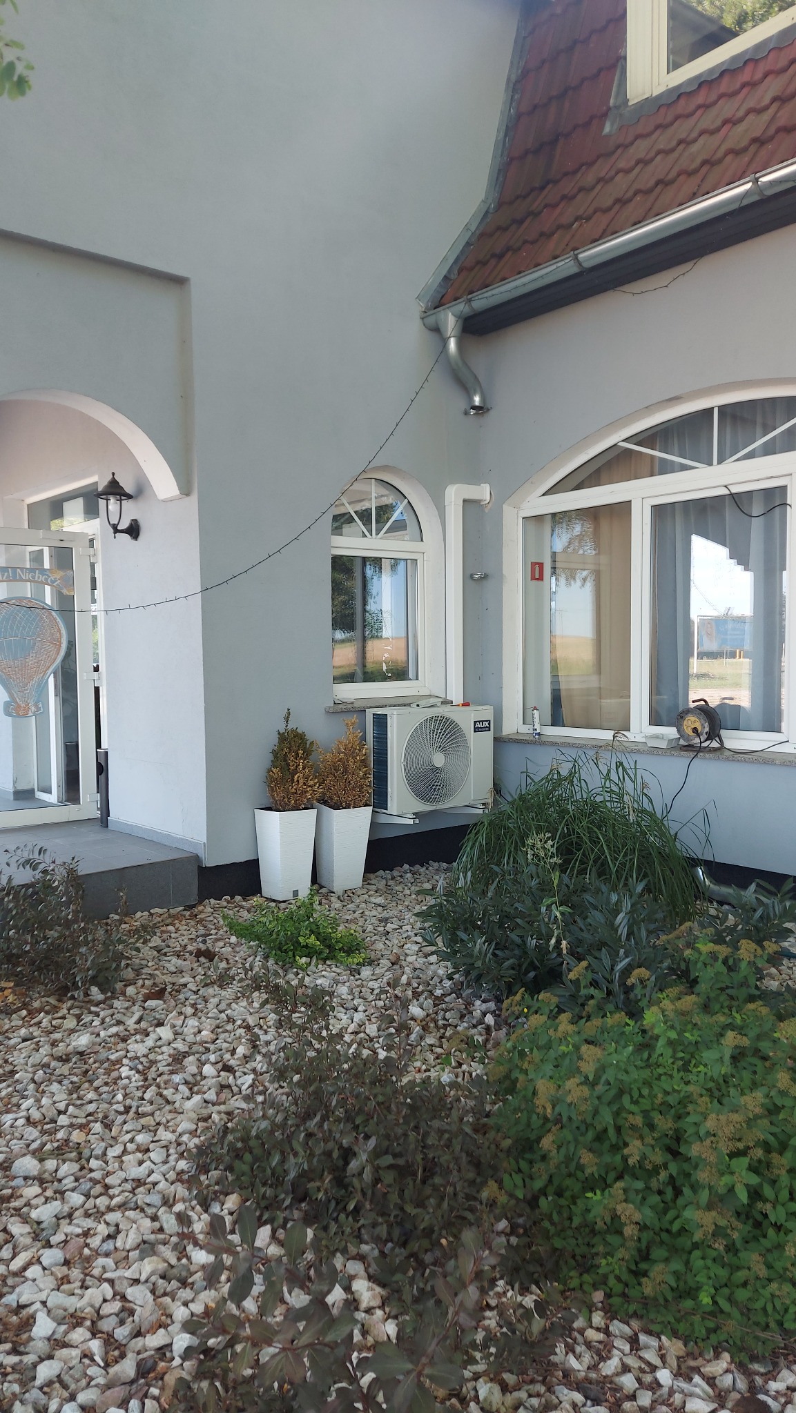 Zewnętrzna jednostka klimatyzacji AUX zamontowana na ścianie szarego budynku, obok okna z białymi ramami, z widocznymi donicami z roślinami i kamieniami ozdobnymi u podstawy.