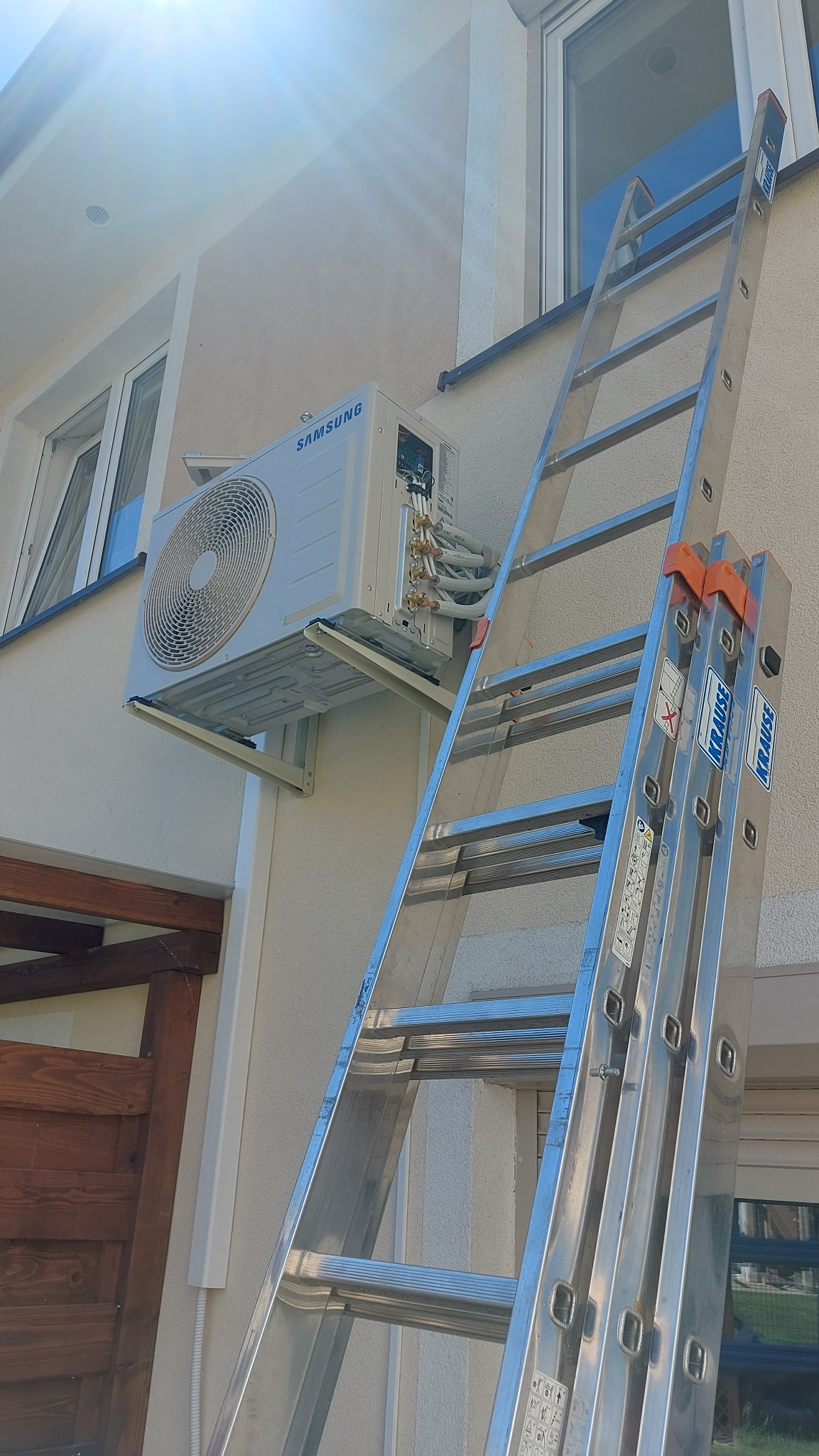 Zewnętrzna jednostka klimatyzacji Samsung zamontowana na ścianie domu, widoczna drabina Krause oparta o ścianę, przewody połączeniowe w trakcie montażu.