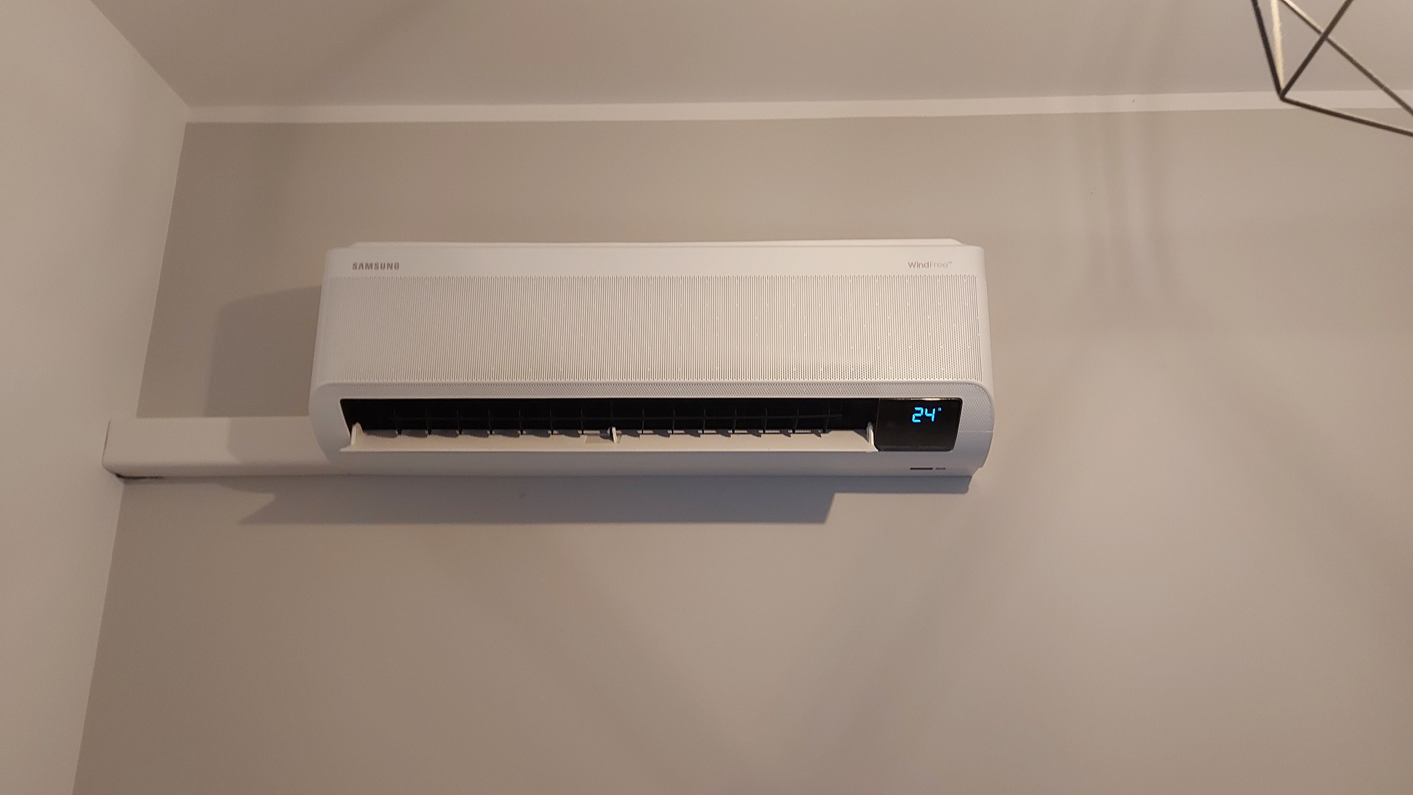 Biały klimatyzator Samsung WindFree zamontowany na szarej ścianie w pomieszczeniu, wyświetlający temperaturę 24 stopnie Celsjusza.