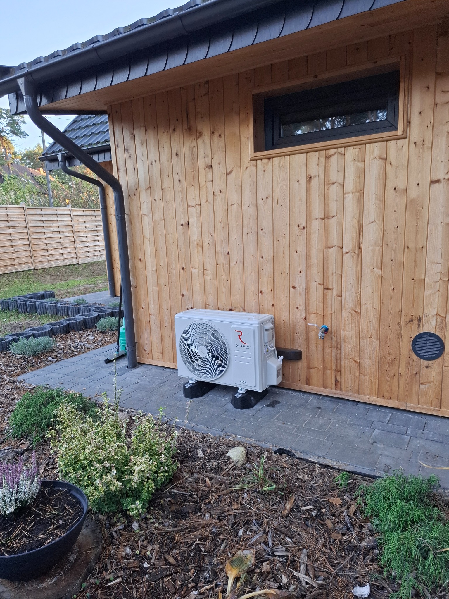 Biała jednostka zewnętrzna klimatyzacji Rotenso zamontowana przy drewnianej ścianie budynku z małym oknem i elementami ogrodu.