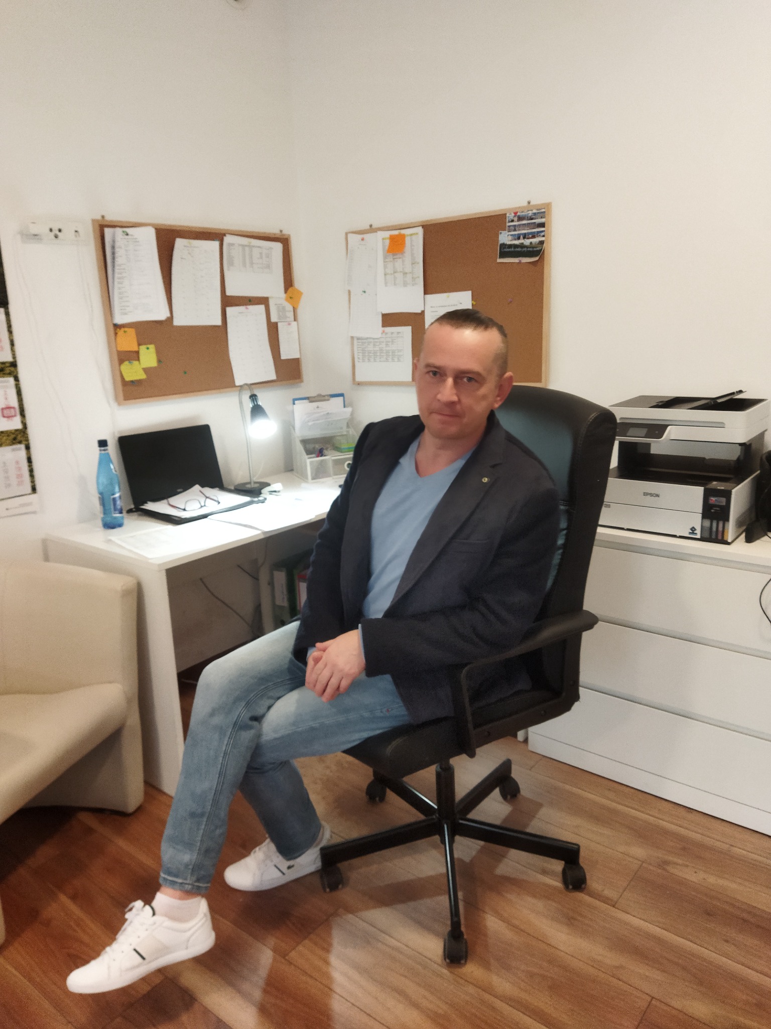 Mężczyzna w niebieskiej koszulce i marynarce siedzi na czarnym krześle biurowym w gabinecie z biurkiem, laptopem, lampką i drukarką.